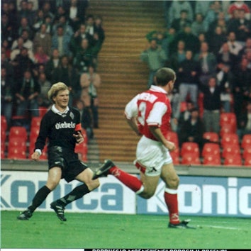 Ein ganz in schwarz gekleideter Fußballer verfolgt gespannt den Flug seines Schusses. Ein Torwart versucht den herannahenden Ball abzuwehren, während ein Verteidiger vergeblich hinterherläuft.