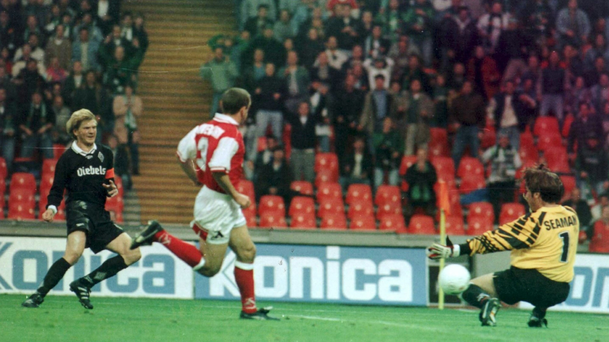 Ein ganz in schwarz gekleideter Fußballer verfolgt gespannt den Flug seines Schusses. Ein Torwart versucht den herannahenden Ball abzuwehren, während ein Verteidiger vergeblich hinterherläuft.