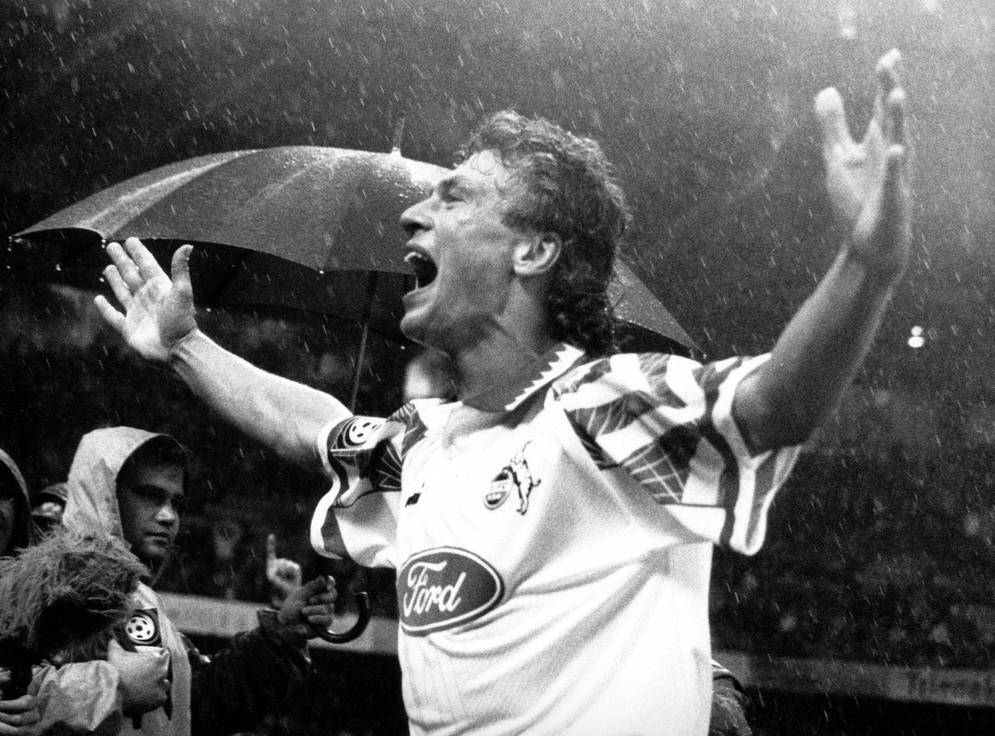 Ein Fußball-Stürmer hat beide Arme zum Jubel ausgestreckt, während im Hintergrund ein Ordner einen Regenschirm hält. Es regnet.