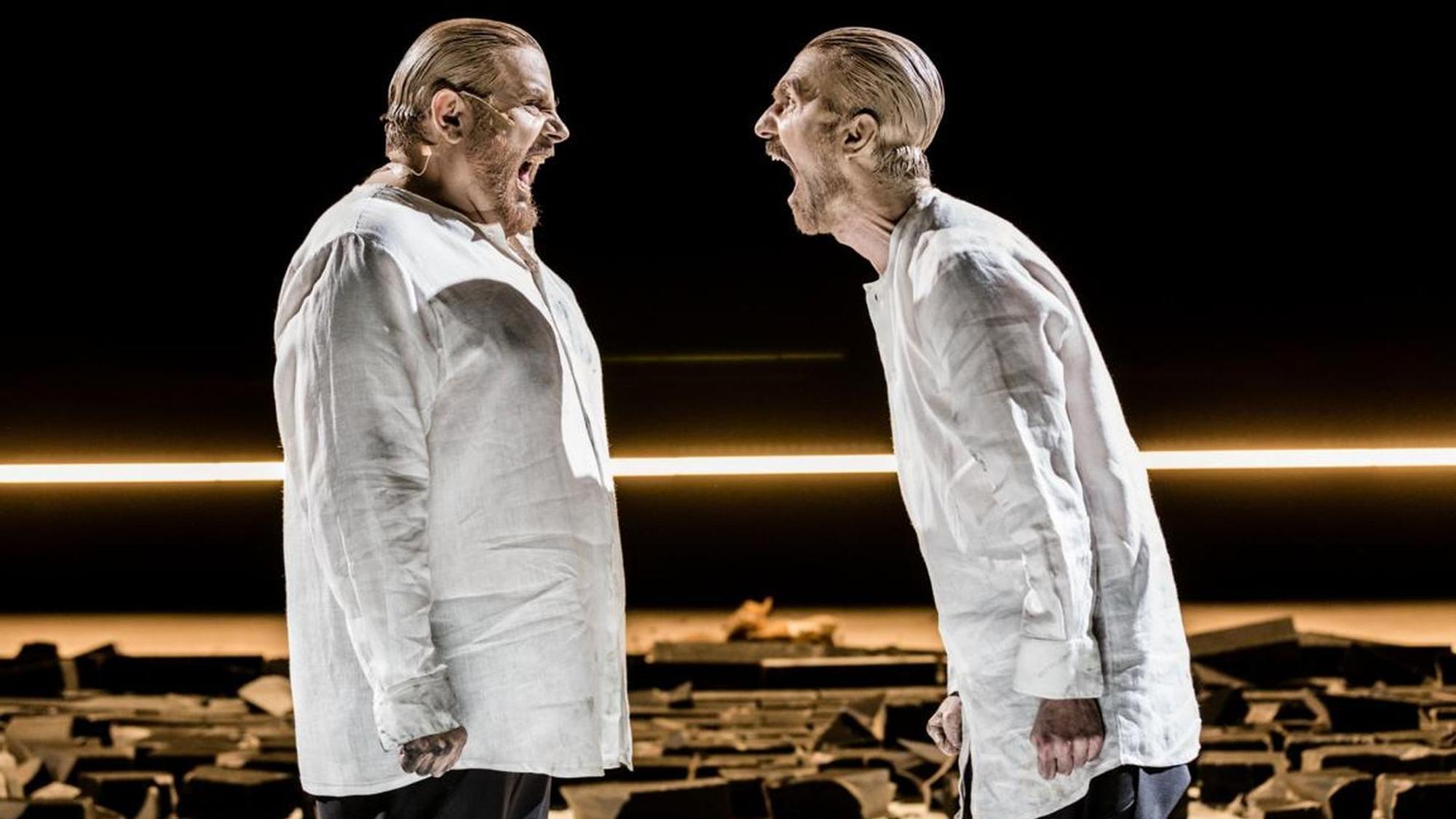 Die Schauspieler Bruno Cathomas (l.) und Seán McDonagh stehen sich gegenüber und schreien sich an. Sie tragen weiße Hemden, ihre Köpfe sind mit Lehm verschmiert.