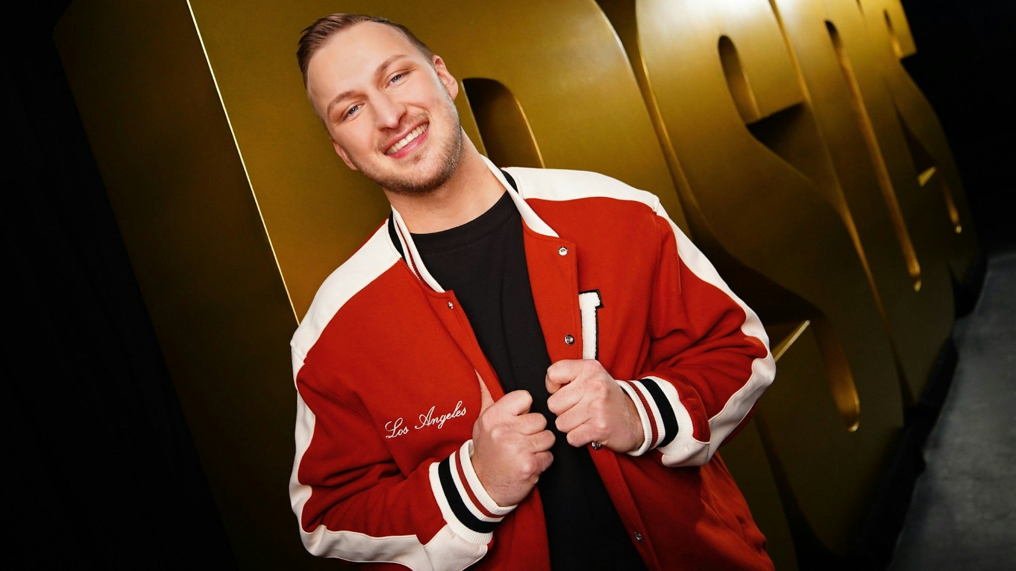 David Leischik steht in einer roten College-Jacke vor „DSDS“-Buchstaben in Gold.