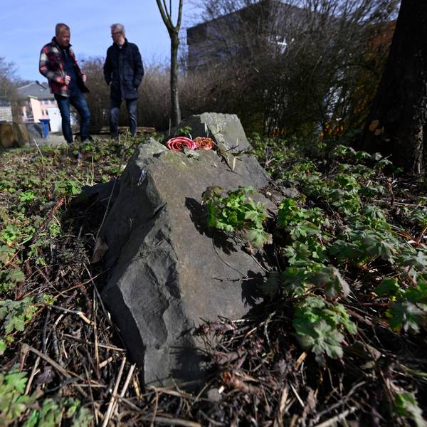 Zwei Männer stehen an einem Gedenkstein, auf dem Rosen liegen.