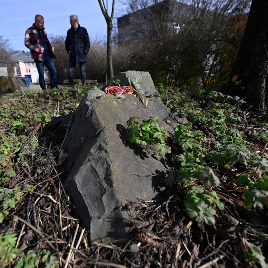 Zwei Männer stehen an einem Gedenkstein, auf dem Rosen liegen.