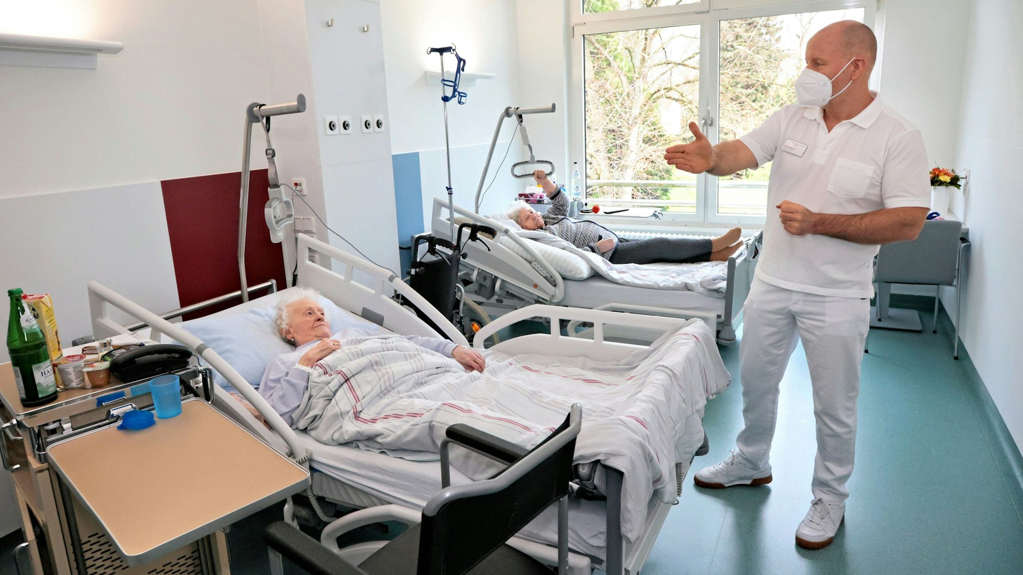 Sascha Wihstutz steht in einem Krankenhauszimmer. Zwei ältere Frauen liegen in ihren Betten.