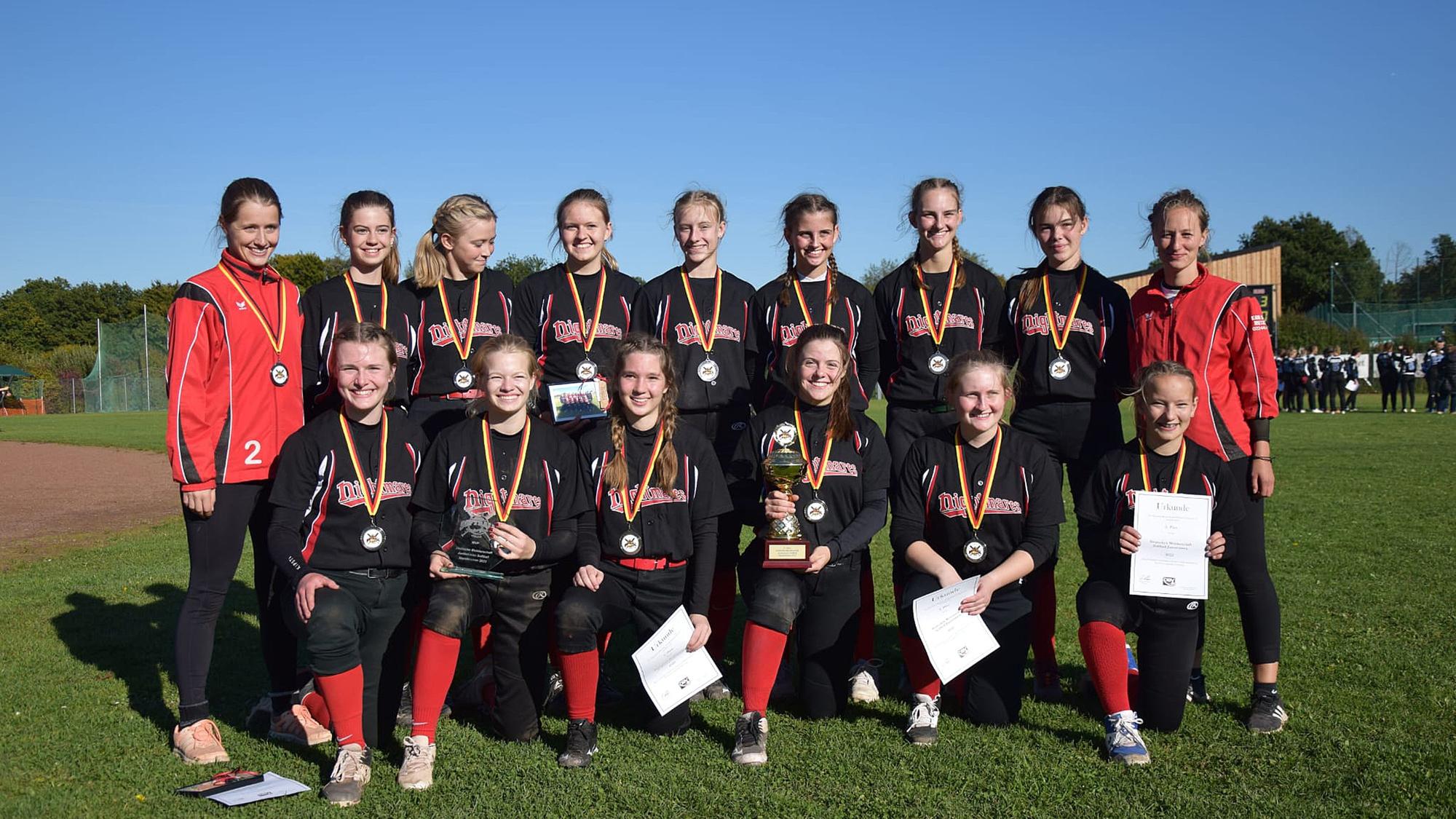 Die Softballerinnen posieren mit umgehangenen Medaillen, Urkunden und dem Pokal für ein Foto.