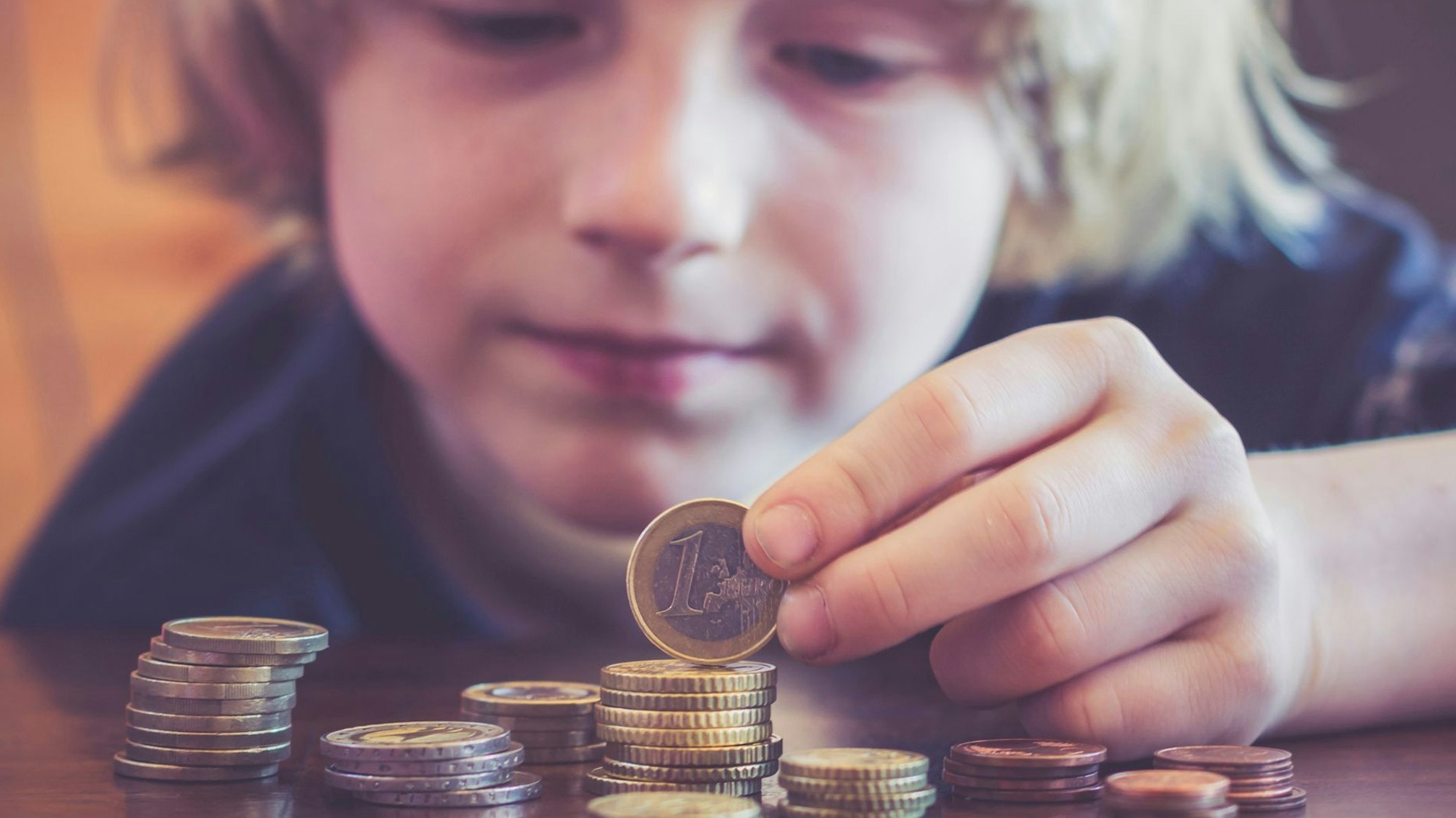 Ein kleiner Junge mit längeren blonden Haaren türmt Münzgeld aufeinander.