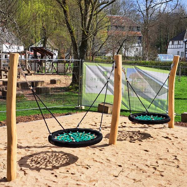 Eine neue Doppelschaukel steht auf einem Sandplatz, noch umgeben von Gitterzäunen. Im Hintergrund sind Fachwerkgebäude des Papiermuseums Alte Dombach zu sehen.