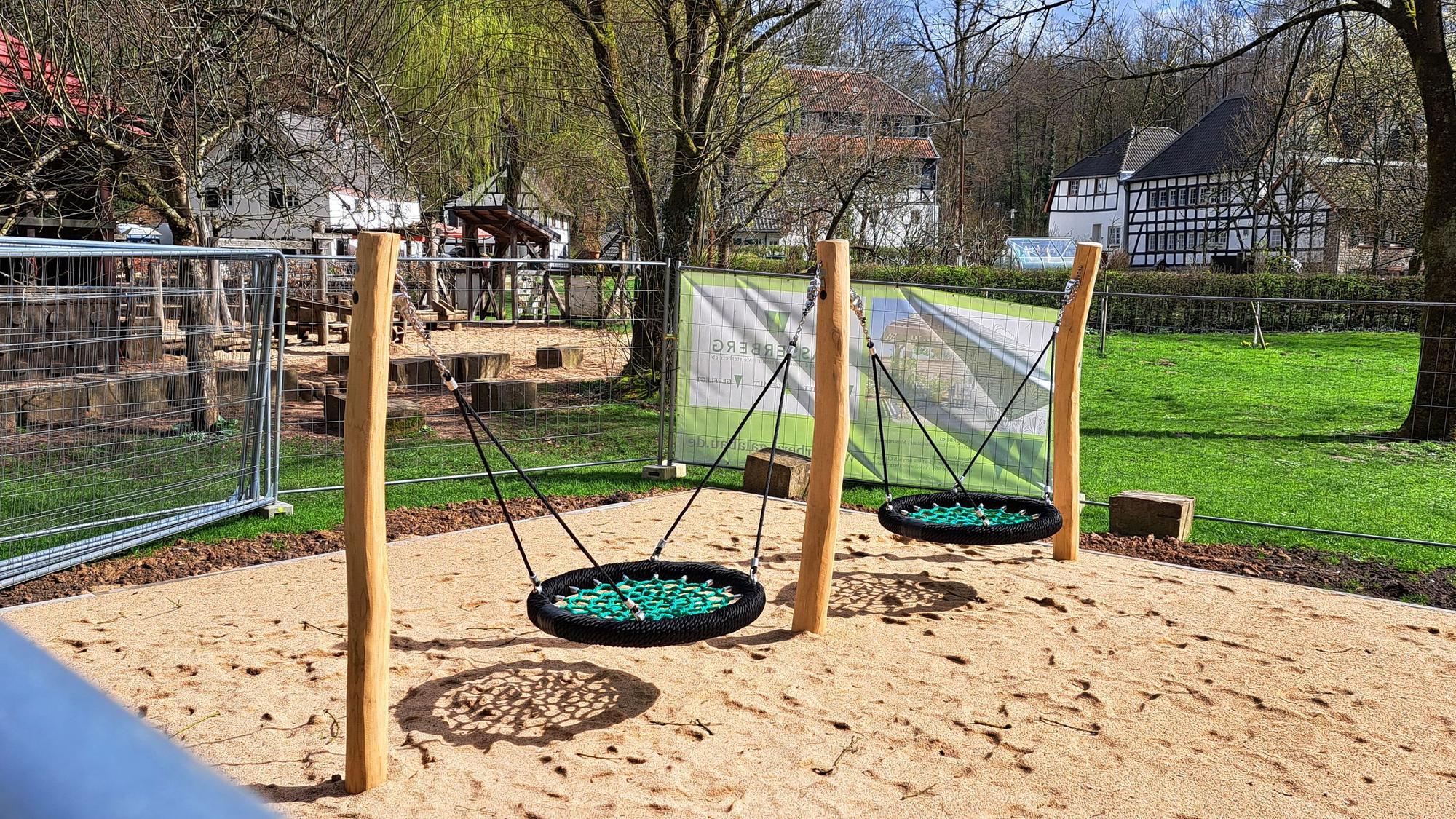 Eine neue Doppelschaukel steht auf einem Sandplatz, noch umgeben von Gitterzäunen. Im Hintergrund sind Fachwerkgebäude des Papiermuseums Alte Dombach zu sehen.