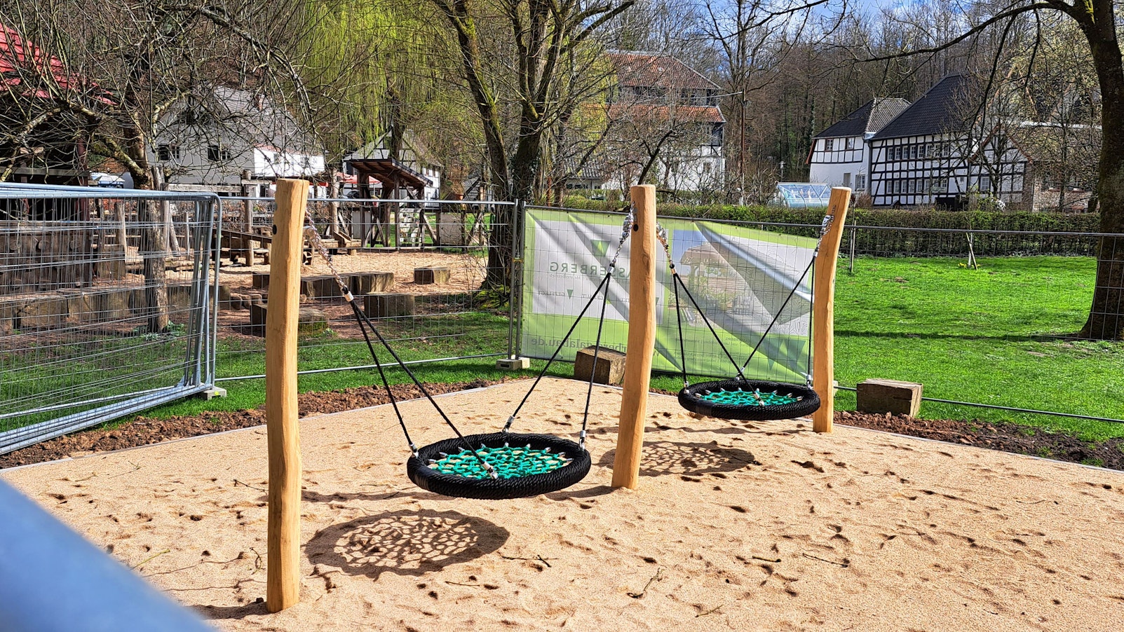 Eine neue Doppelschaukel steht auf einem Sandplatz, noch umgeben von Gitterzäunen. Im Hintergrund sind Fachwerkgebäude des Papiermuseums Alte Dombach zu sehen.