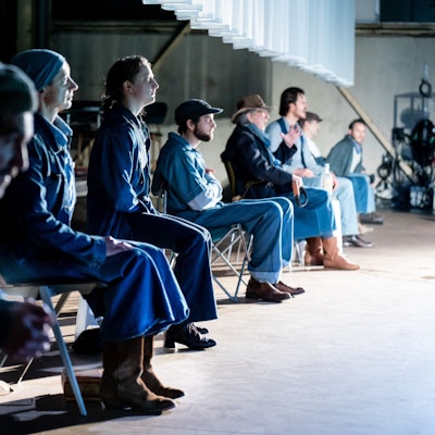 Acht Schauspieler des Kölner Ensembles sitzen, von der Seite fotografiert, nebeneinander auf Klappstühlen im Stück „Früchte des Zorns“ im Depot 2. Sie tragen alle Kostüme aus Jeansstoff.