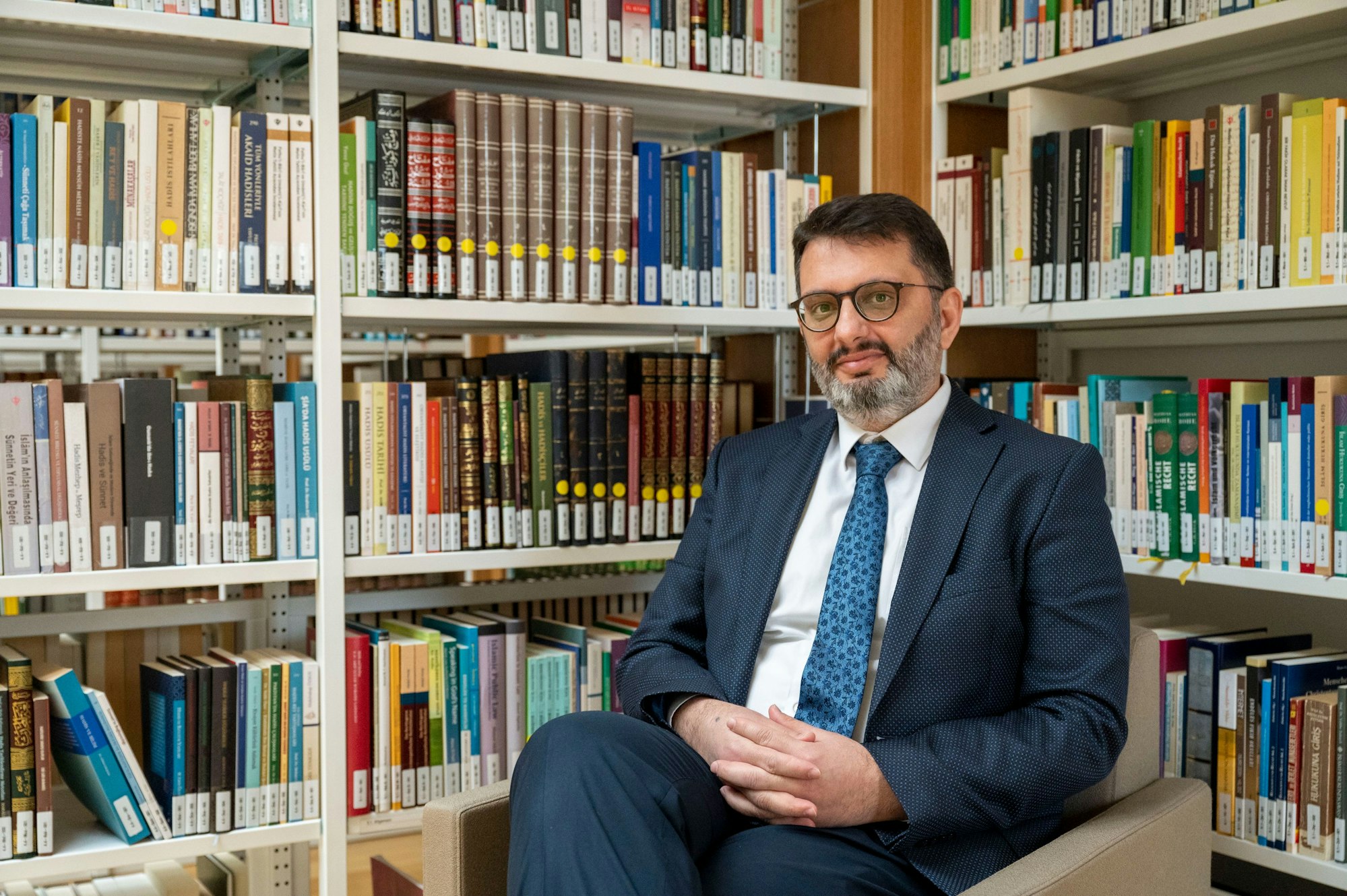 Muharrem Kuzey sitzt auf einem Sessel in der Bibliothek der Zentralmoschee. Er trägt einen Anzug, um ihn herum stehen Bücherregale.