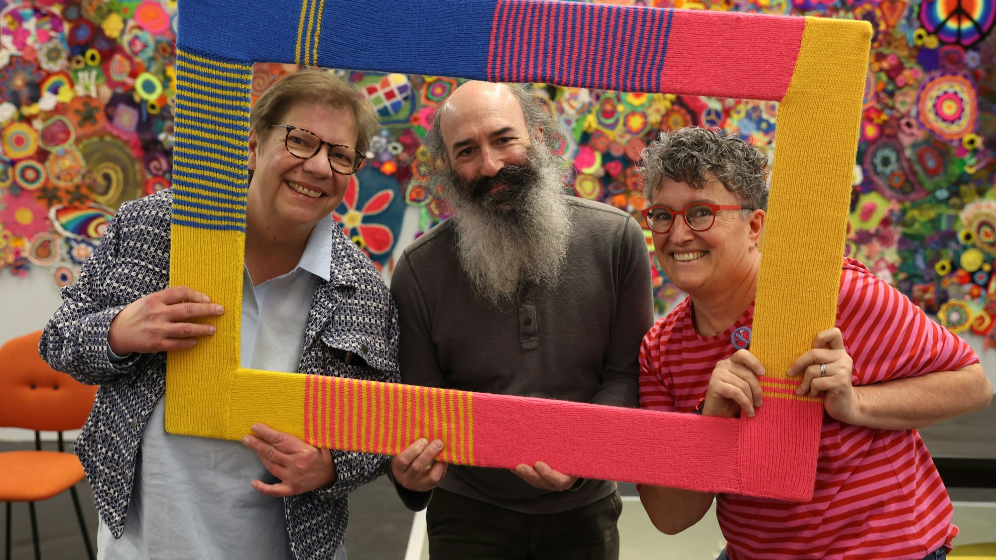 Die drei halten einen gehäkelten Rahmen, die Peace-Zeichen im Hintergrund