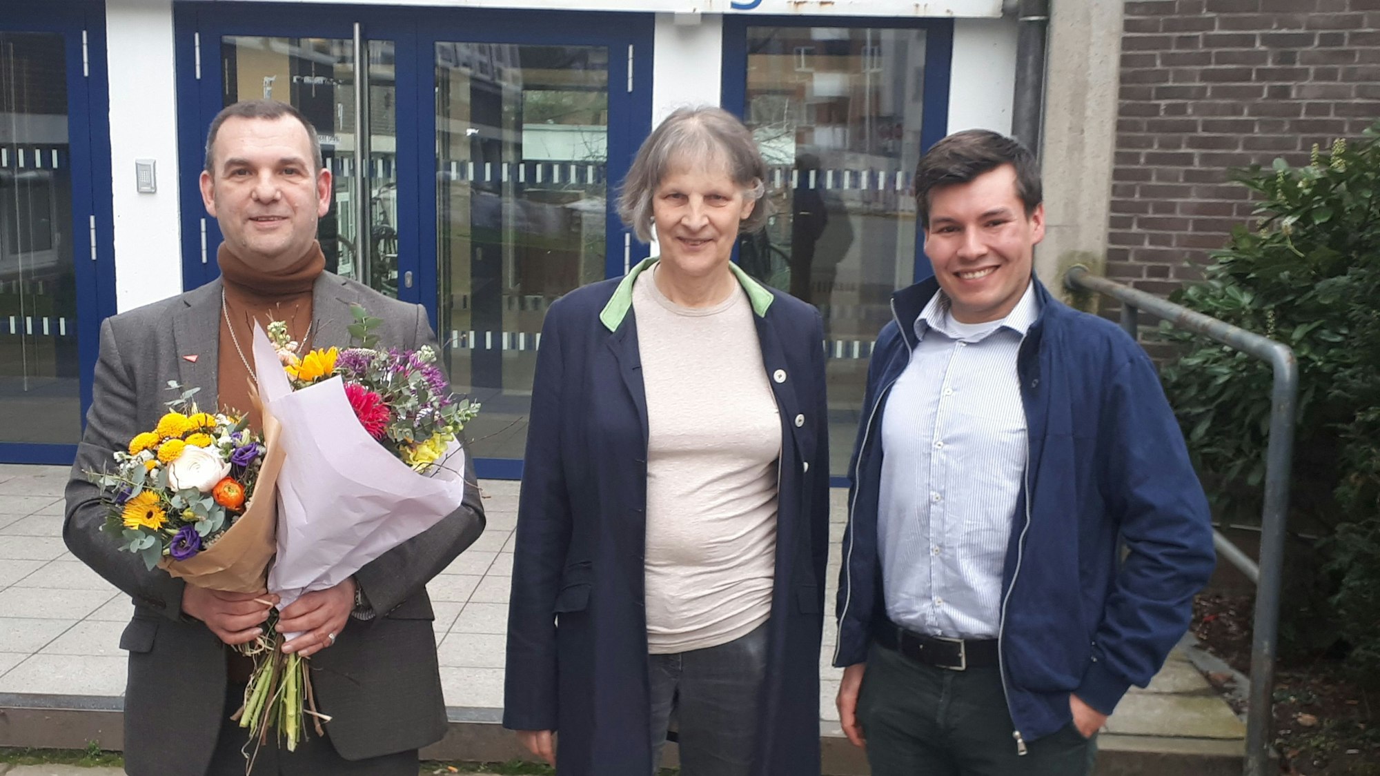 Drei Politiker stehen vor der Schule, der neu Gewählte hat Blumen in der Hand.