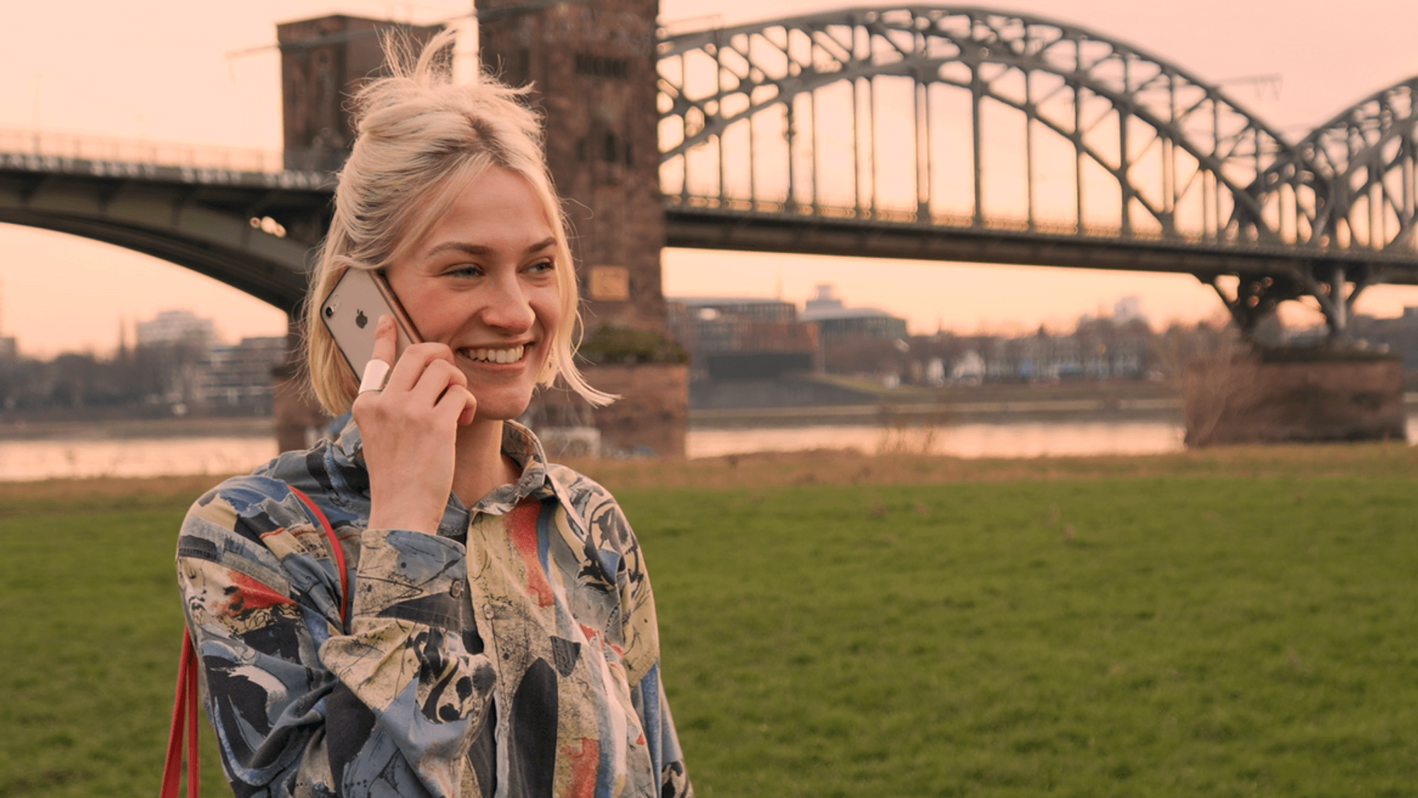 Eine junge Frau steht vor einer Kölner Brücke und telefoniert.