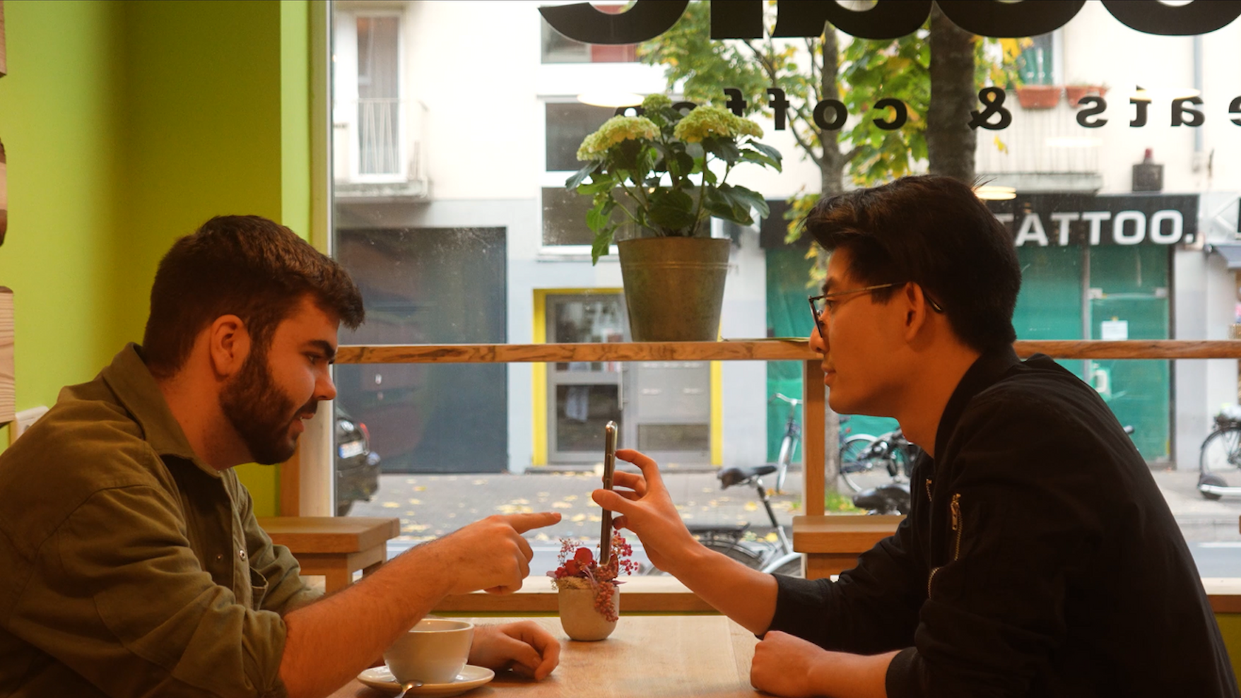 Zwei junge Männer sitzen in einem Kaffee und halten ein Smartphone hoch.