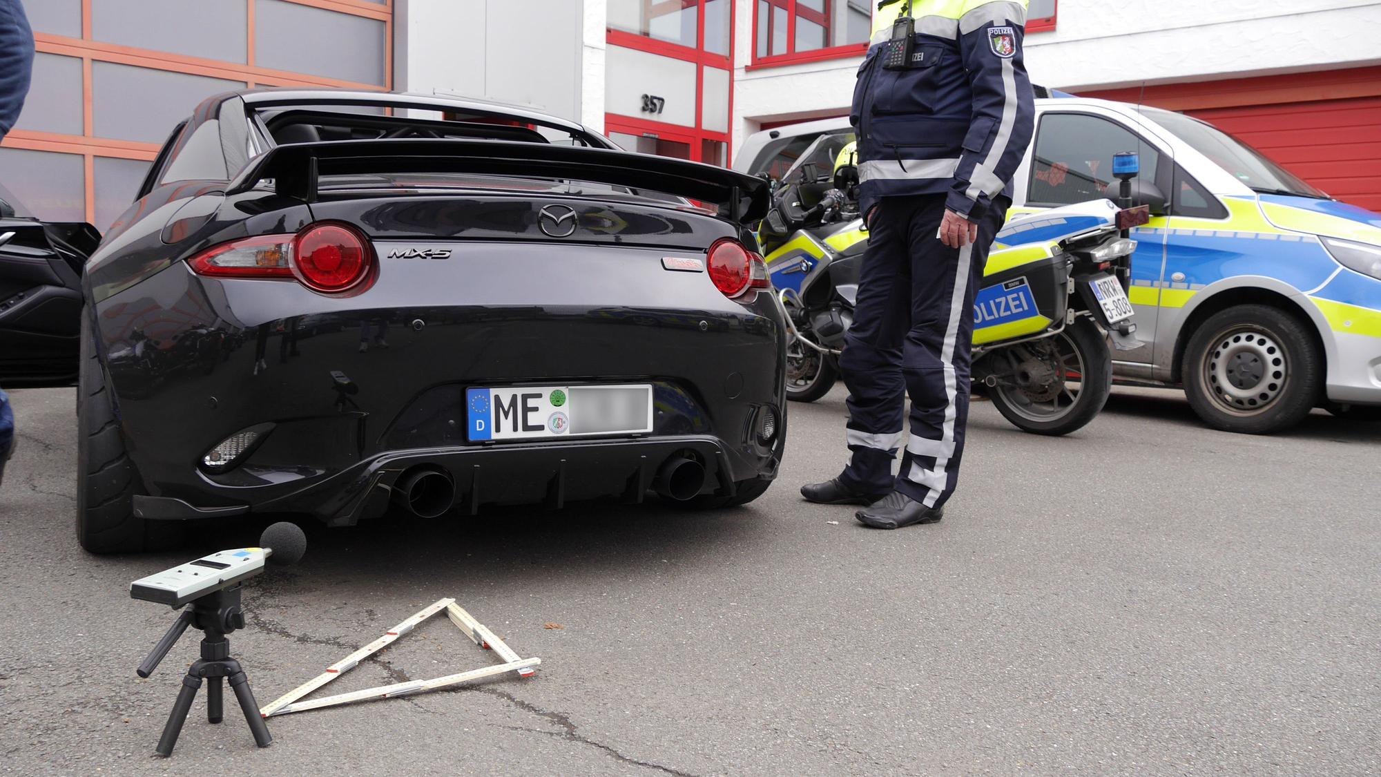 Hinter dem Auspuff eines Autos steh ein Messgerät auf dem Boden. Im Hintergrund parken ein Polizeimotorrad und ein Streifenwagen.