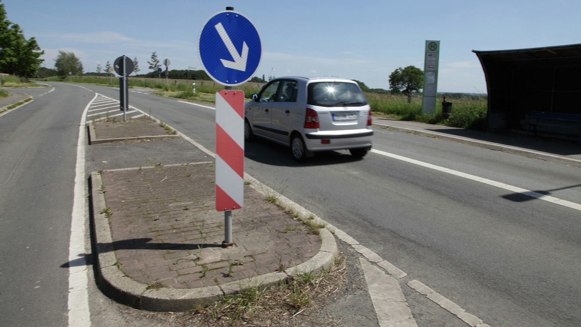 An einer Querungshilfe auf einer zweispurigen Straße fährt ein Auto vorbei. Rechts im Bild: eine Bushaltestelle.