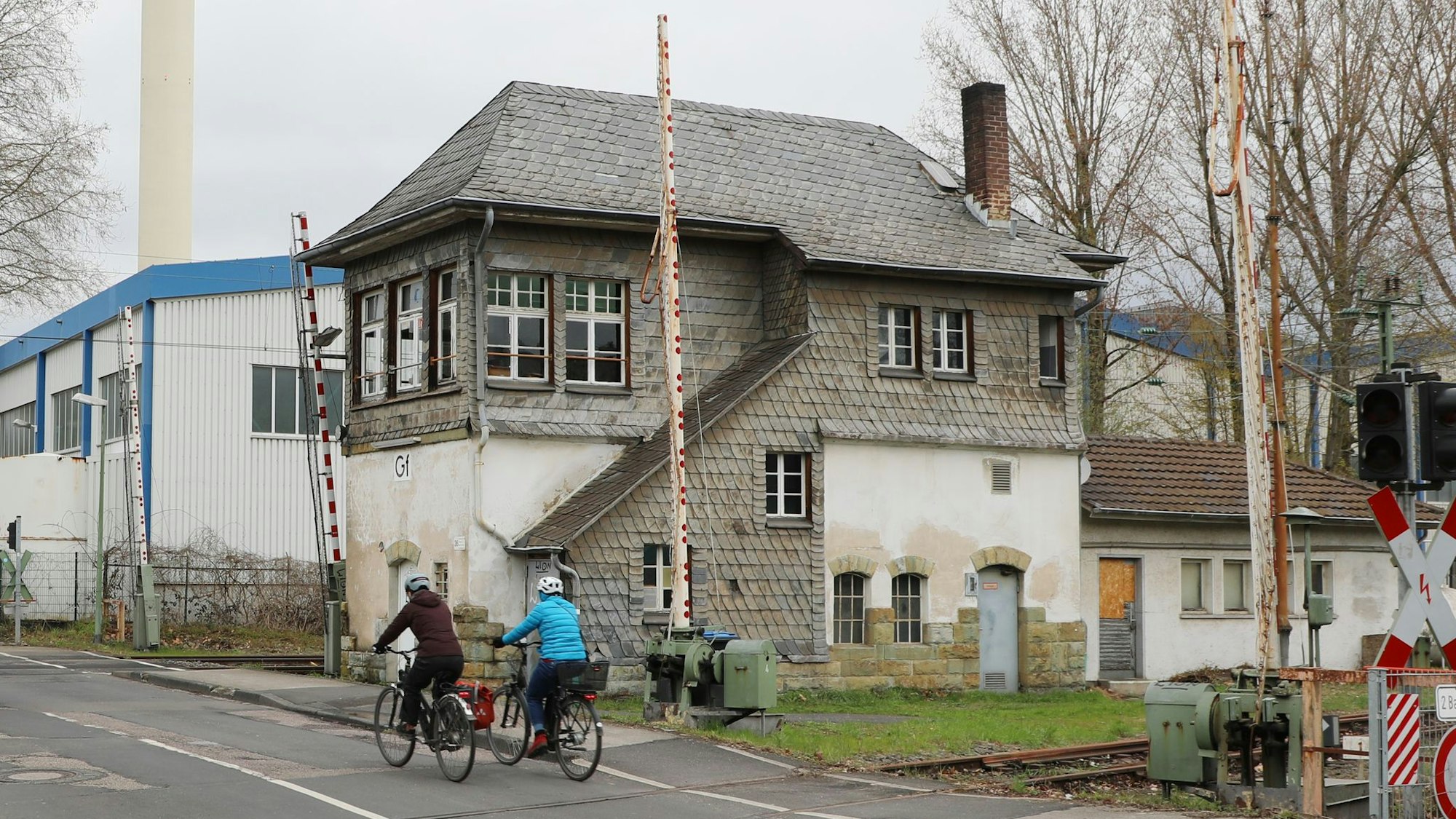 Zwei Radfahrer fahren über einen Bahnübergang, an dem ein historisches Stellwerkgebäude steht. Links und rechts des Stellwerks sind Schrankenanlagen zu sehen.
