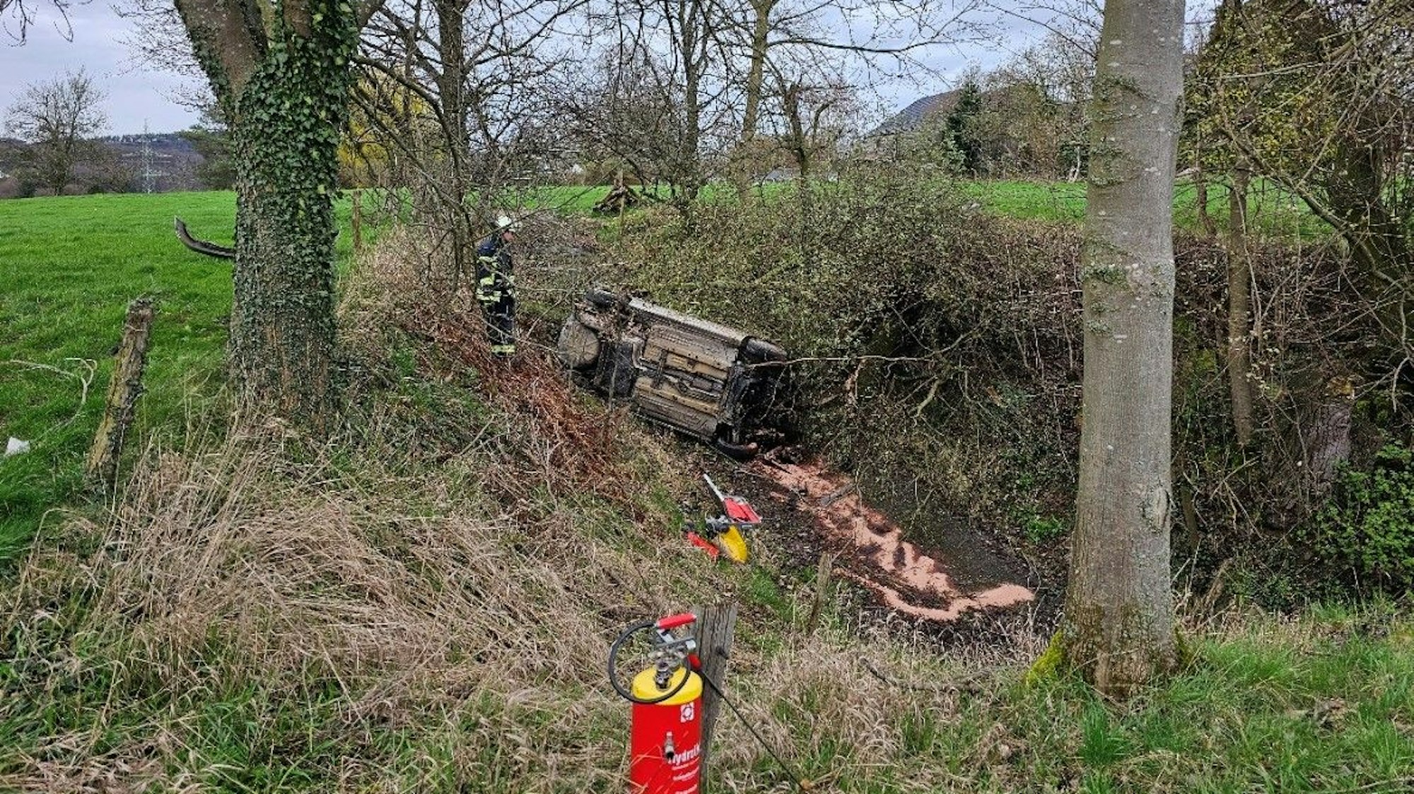 Ein Auto liegt auf der Seite in einer Böschung. Daneben steht ein Feuerwehrmann. Im Vordergrund steht ein Feuerwehrlöscher an einem Zaunpfahl.