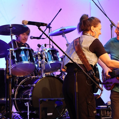 Zwei Musiker, eine Frau und ein Mann, mit Gitarre, stehen zueinander gewandt auf einer Bühne. Im Hintergrund ist ein Schlagzeuger zu sehen.