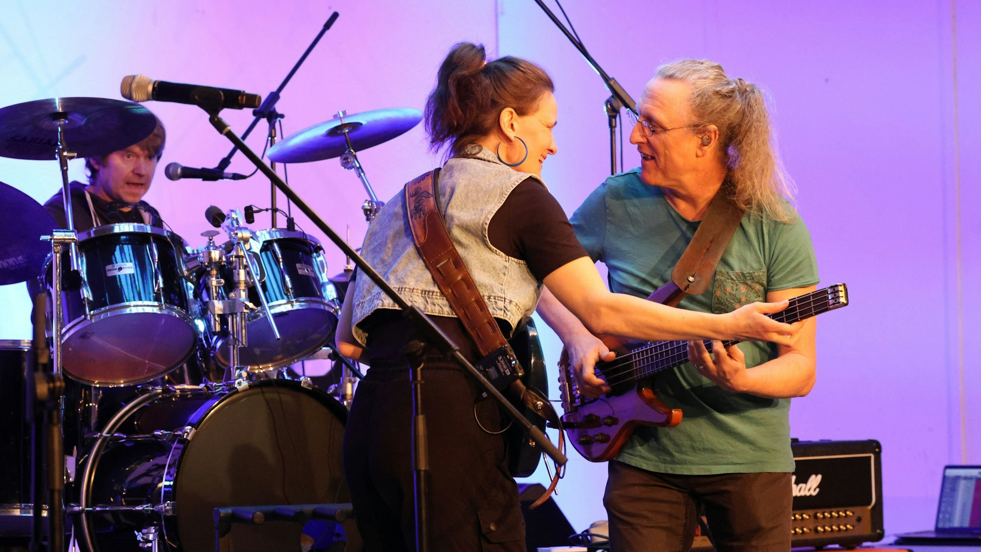 Zwei Musiker, eine Frau und ein Mann, mit Gitarre, stehen zueinander gewandt auf einer Bühne. Im Hintergrund ist ein Schlagzeuger zu sehen.