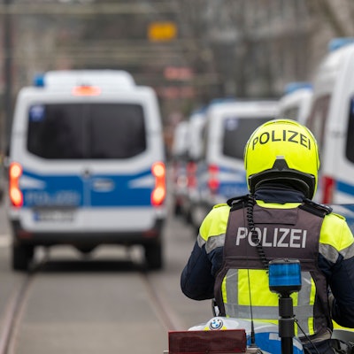 Zu sehen ist ein Polizist auf einem Motorrad in der Rückansicht. Davor befinden sich mehrere Einsatzfahrzeuge der Polizei.