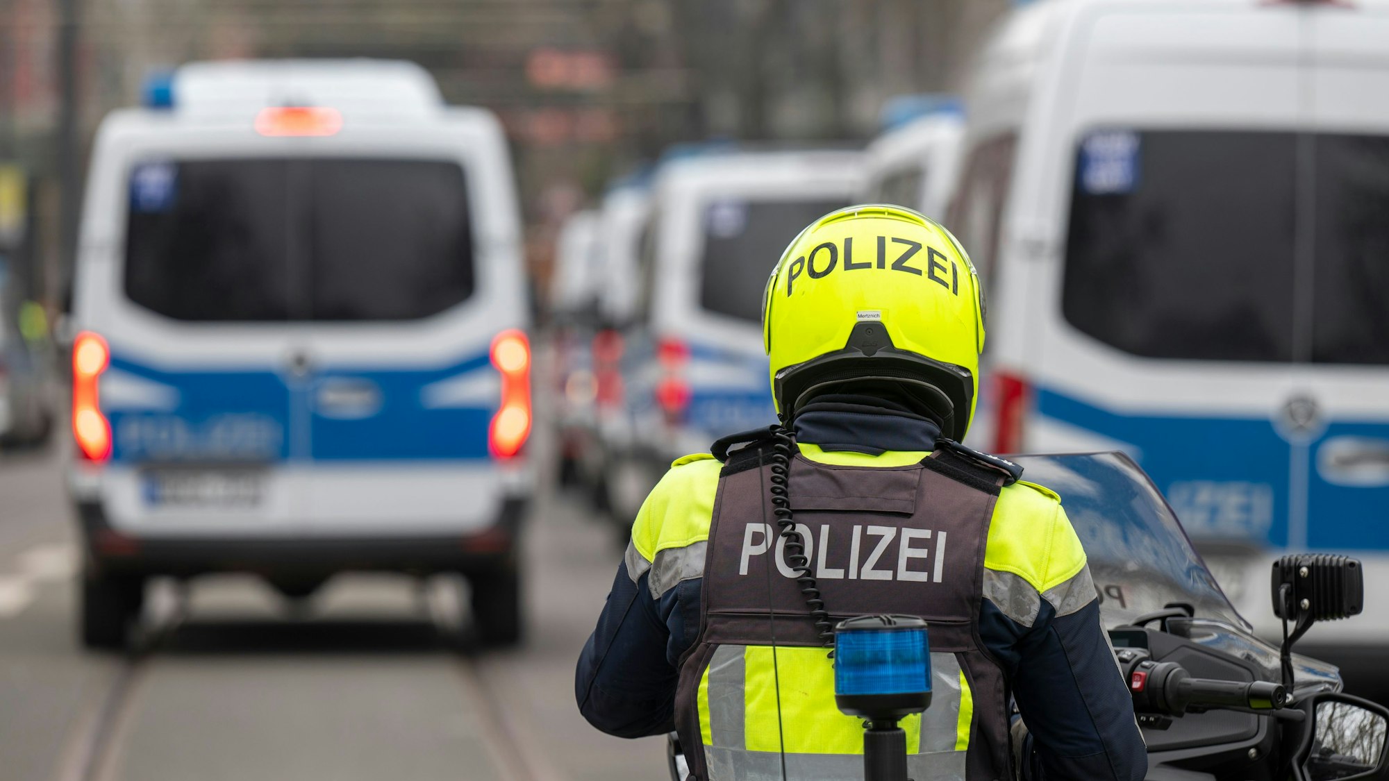 Zu sehen ist ein Polizist auf einem Motorrad in der Rückansicht. Davor befinden sich mehrere Einsatzfahrzeuge der Polizei.