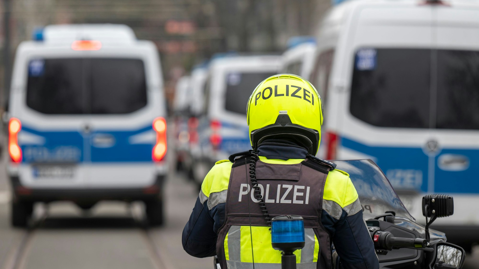 Zu sehen ist ein Polizist auf einem Motorrad in der Rückansicht. Vor ihm befinden sich links und rechts mehrere Einsatzfahrzeuge der Polizei.