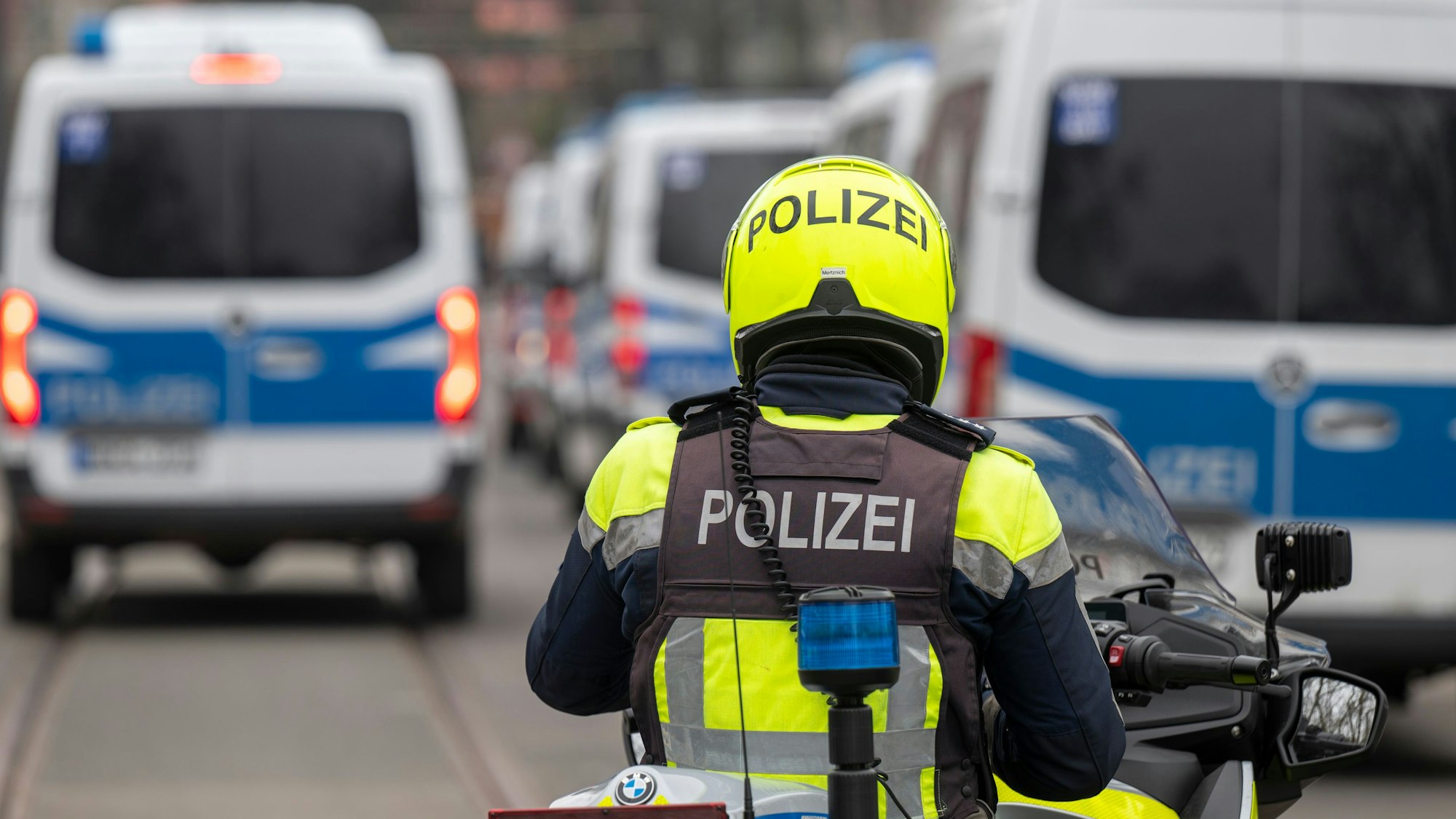 Zu sehen ist ein Polizist auf einem Motorrad in der Rückansicht. Vor ihm befinden sich links und rechts mehrere Einsatzfahrzeuge der Polizei.