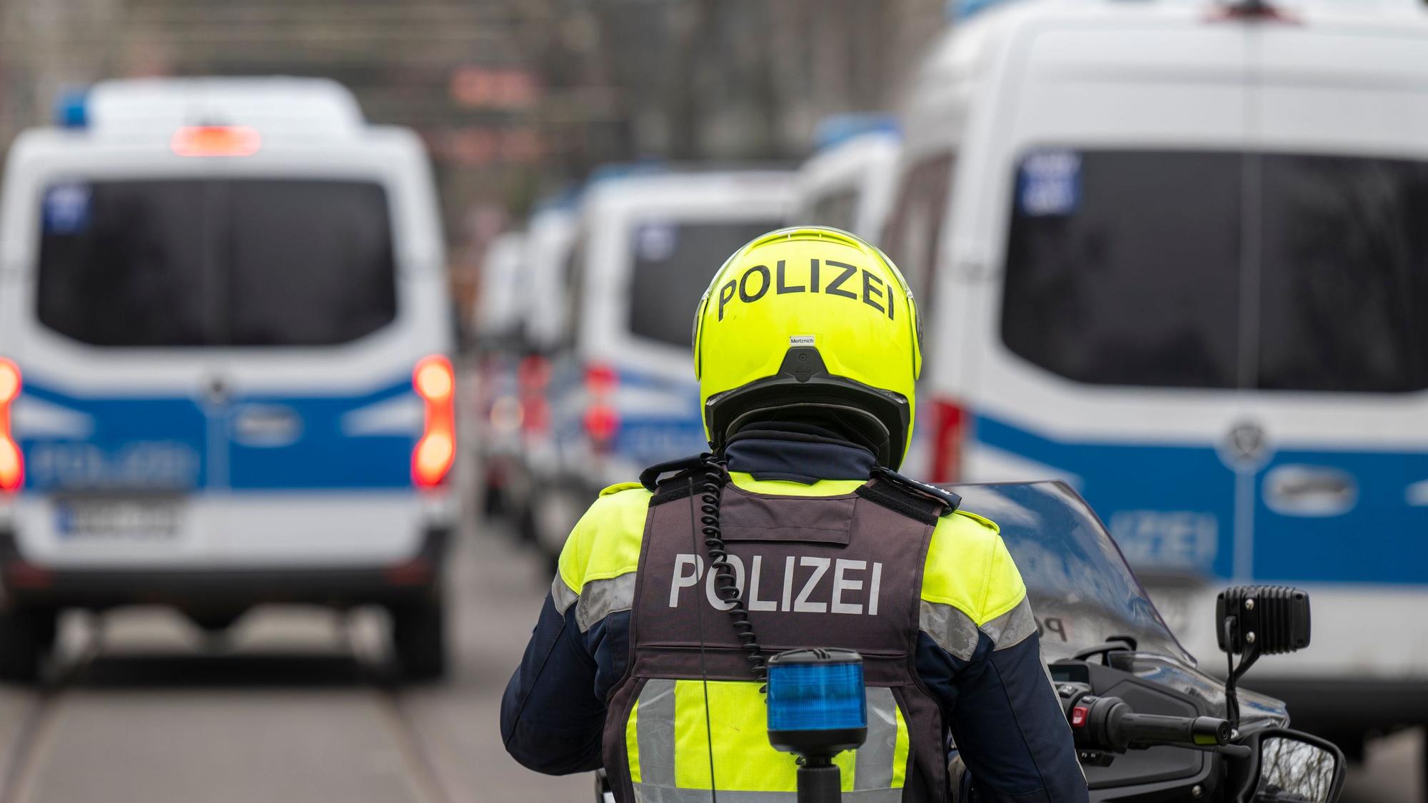 Zu sehen ist ein Polizist auf einem Motorrad in der Rückansicht. Davor sind mehrere Mannschaftswagen der Polizei zu sehen.