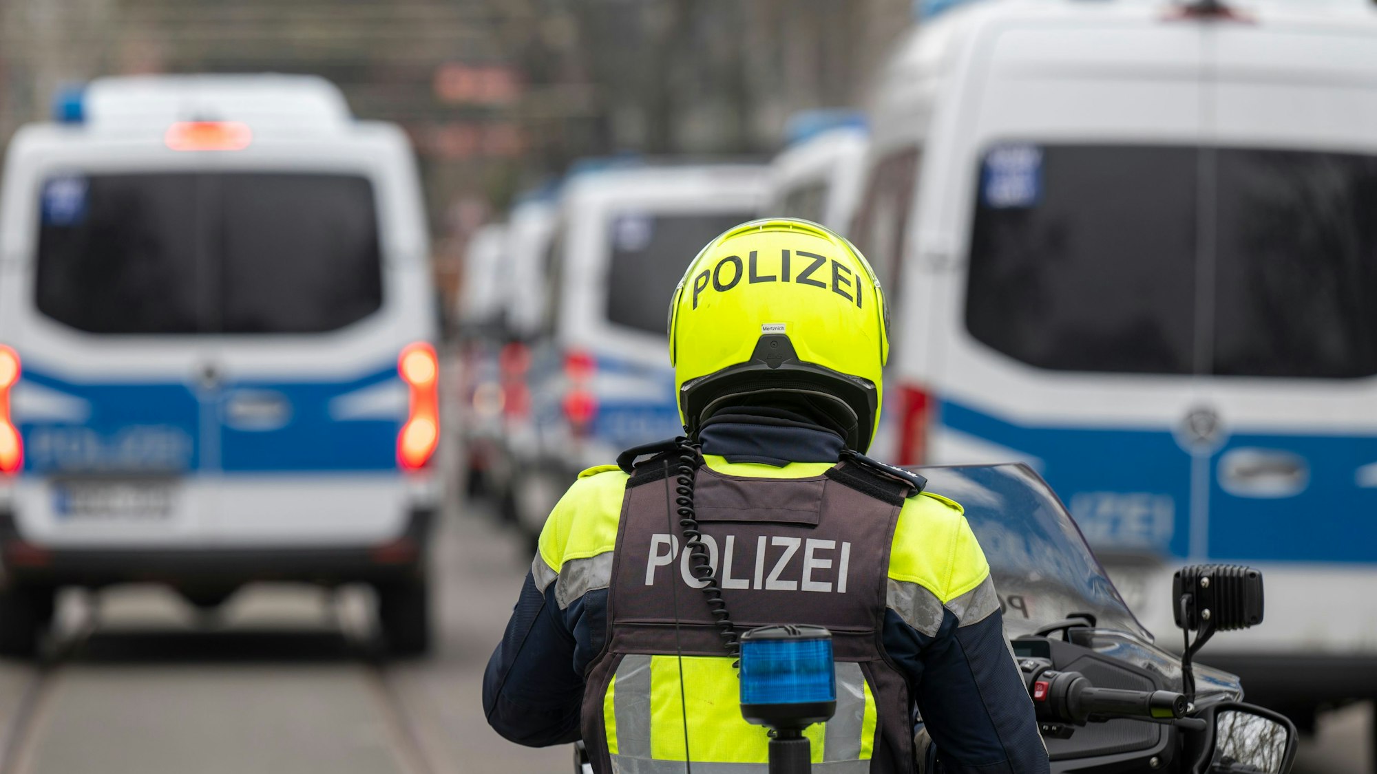 Zu sehen ist ein Polizist auf einem Polizeimotorrad in der Rückansicht. Vor ihm befinden sich links und rechts mehrere Mannschaftswagen der Polizei.