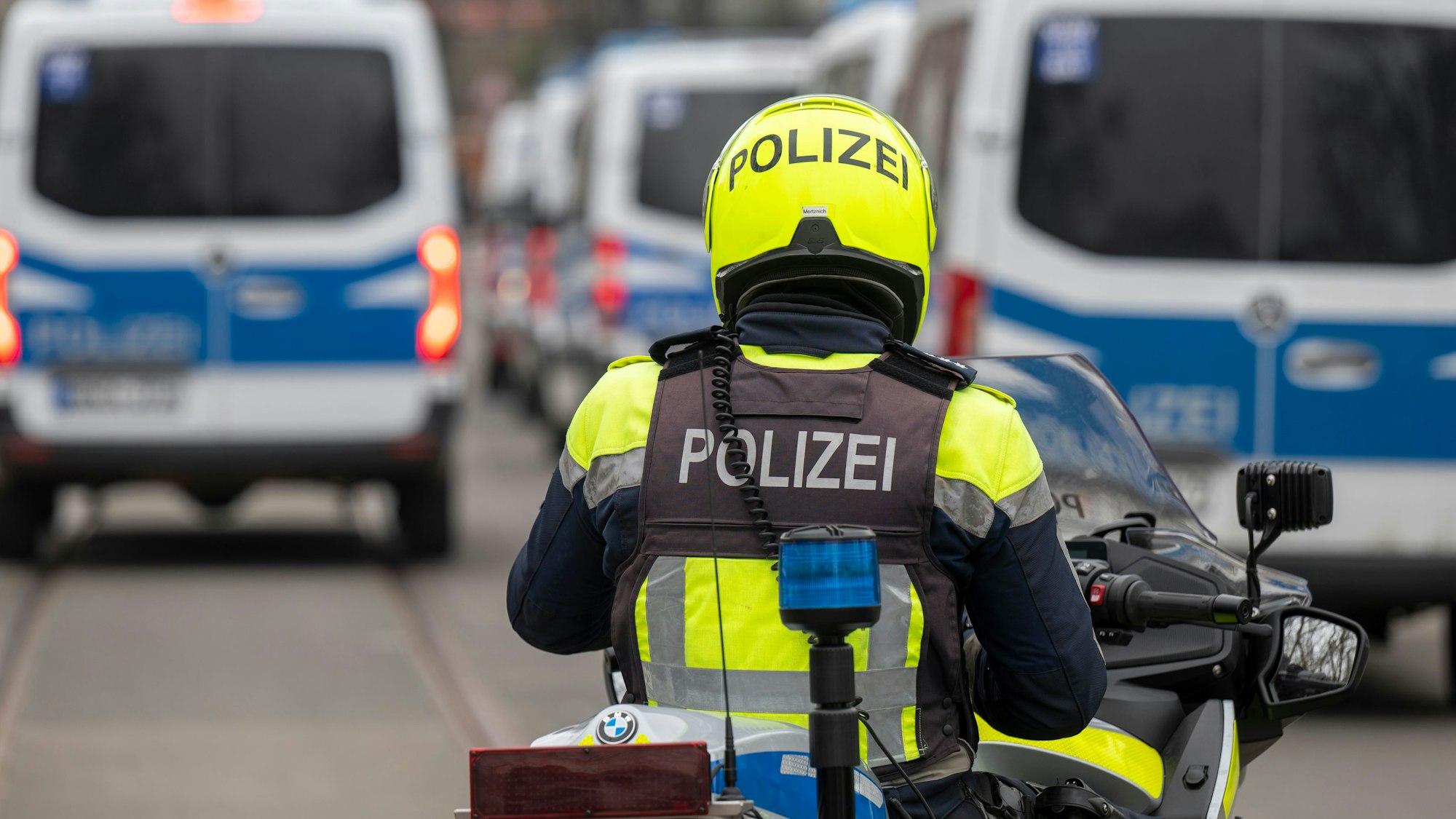 Zu sehen ist ein Polizist auf einem Motorrad in der Rückansicht. Rechts und links vor ihm befinden sich mehrere Mannschaftswagen der Polizei.