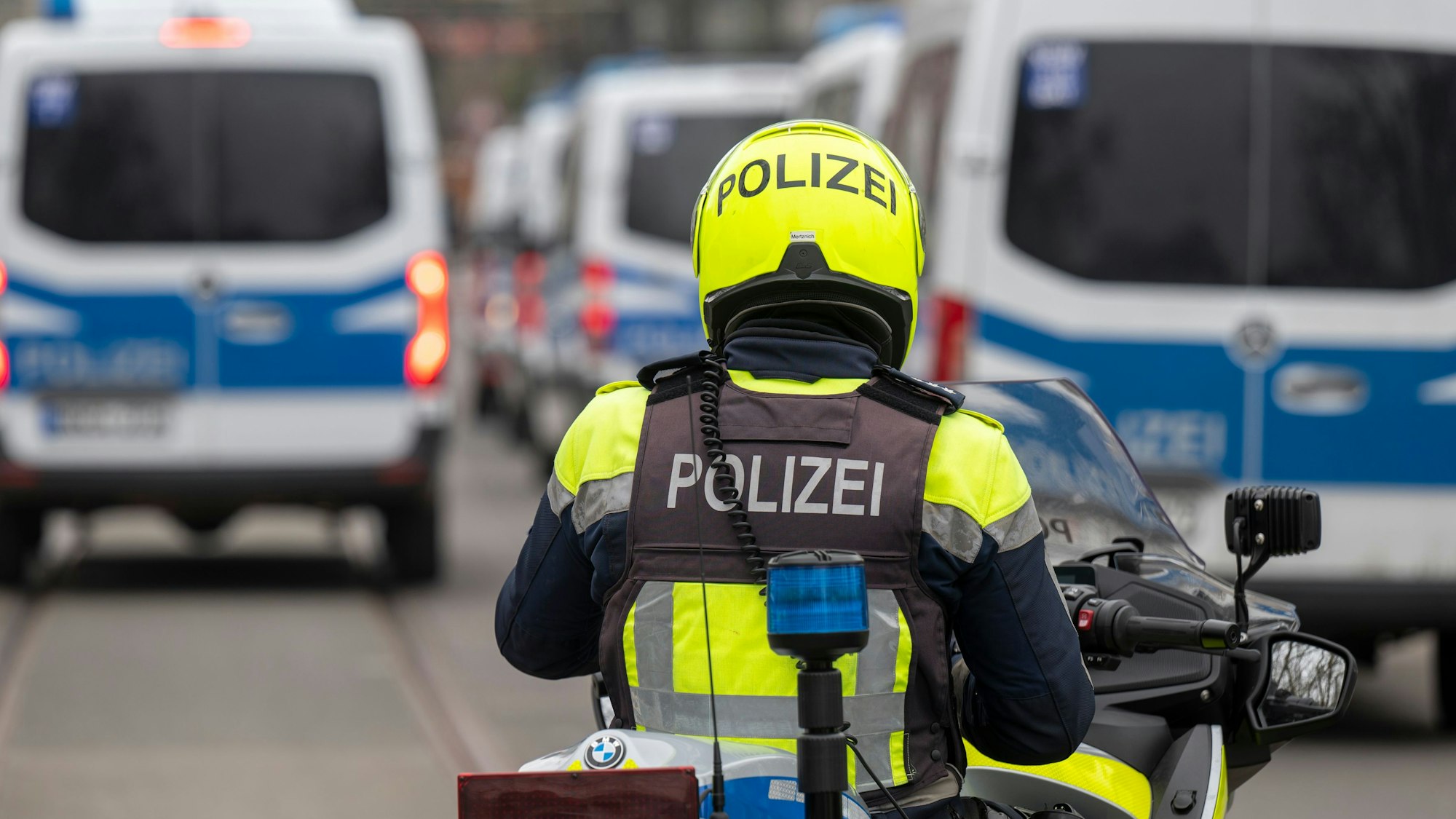 Zu sehen ist ein Polizist auf einem Motorrad in der Rückansicht. Vor ihm befinden sich links und rechts mehrere Mannschaftswagen der Polizei.