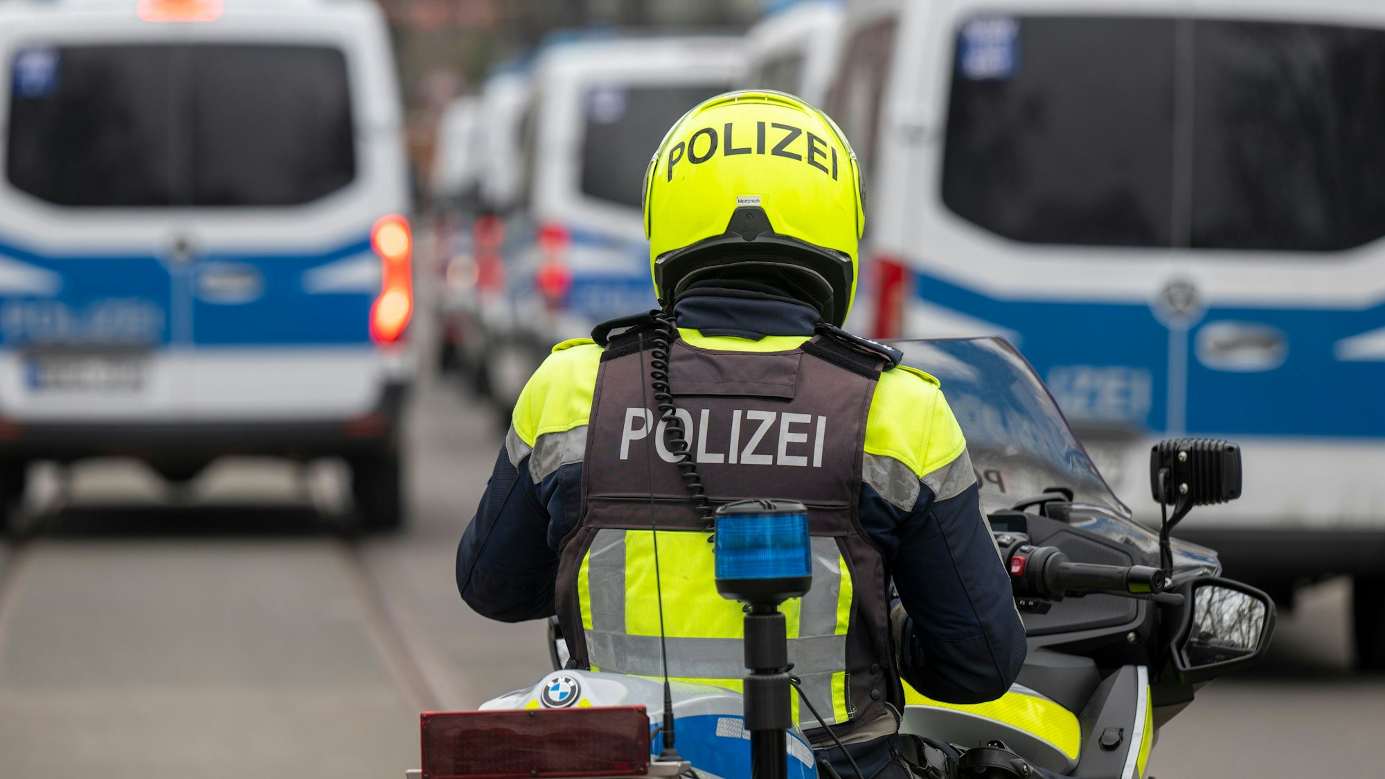 Zu sehen ist ein Polizist auf einem Motorrad in der Rückansicht. Vor ihm befinden sich links und rechts mehrere Einsatzfahrzeuge der Polizei.