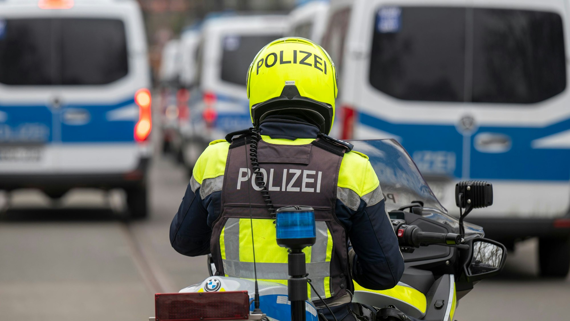 Zu sehen ist ein Polizist auf einem Motorrad in der Rückansicht. Vor ihm befinden sich links und rechts mehrere Einsatzfahrzeuge der Polizei.