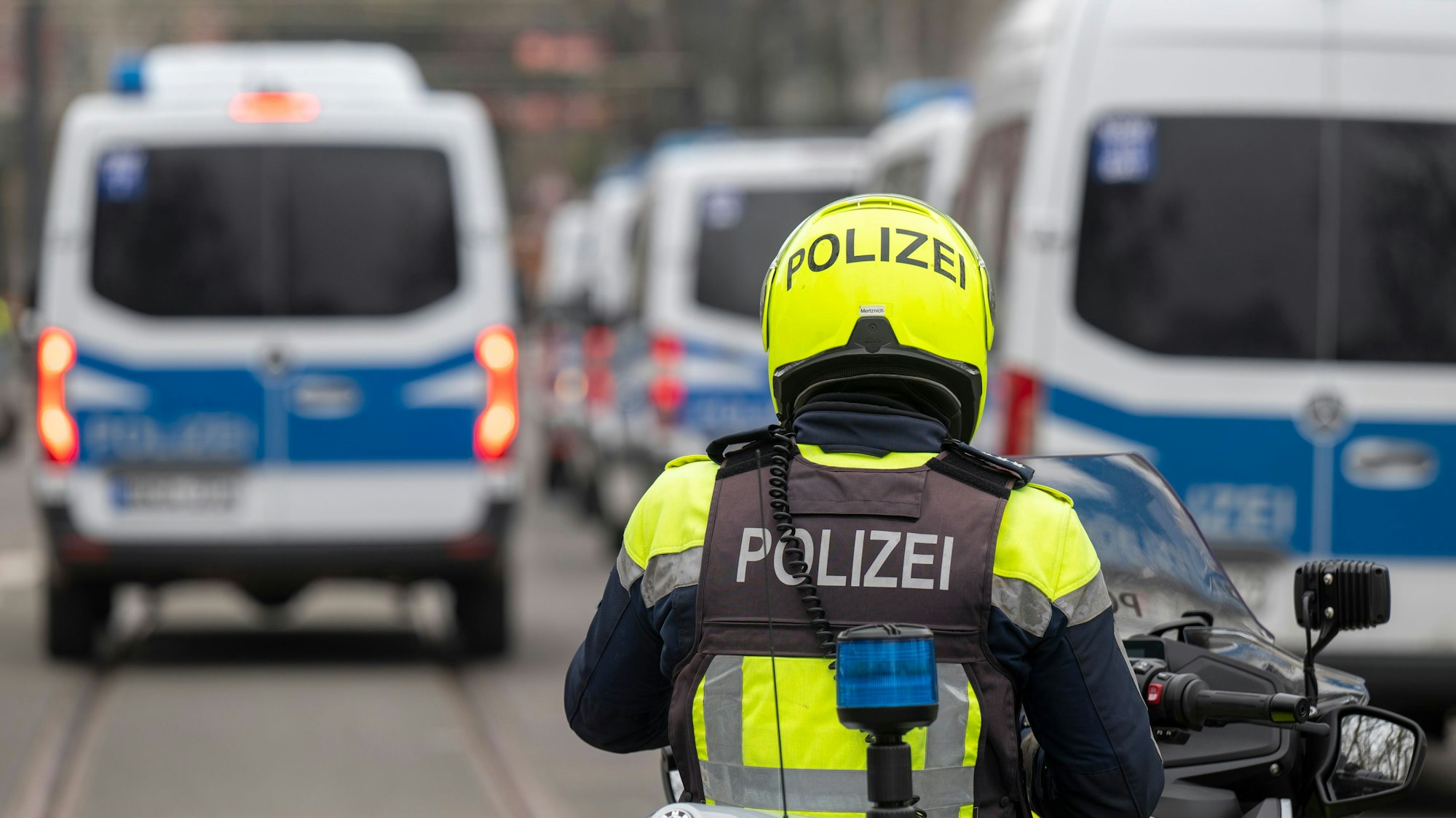 Zu sehen ist ein Polizist auf einem Motorrad in der Rückansicht. Vor ihm befinden sich links und rechts mehrere Transporter der Kölner Polizei.