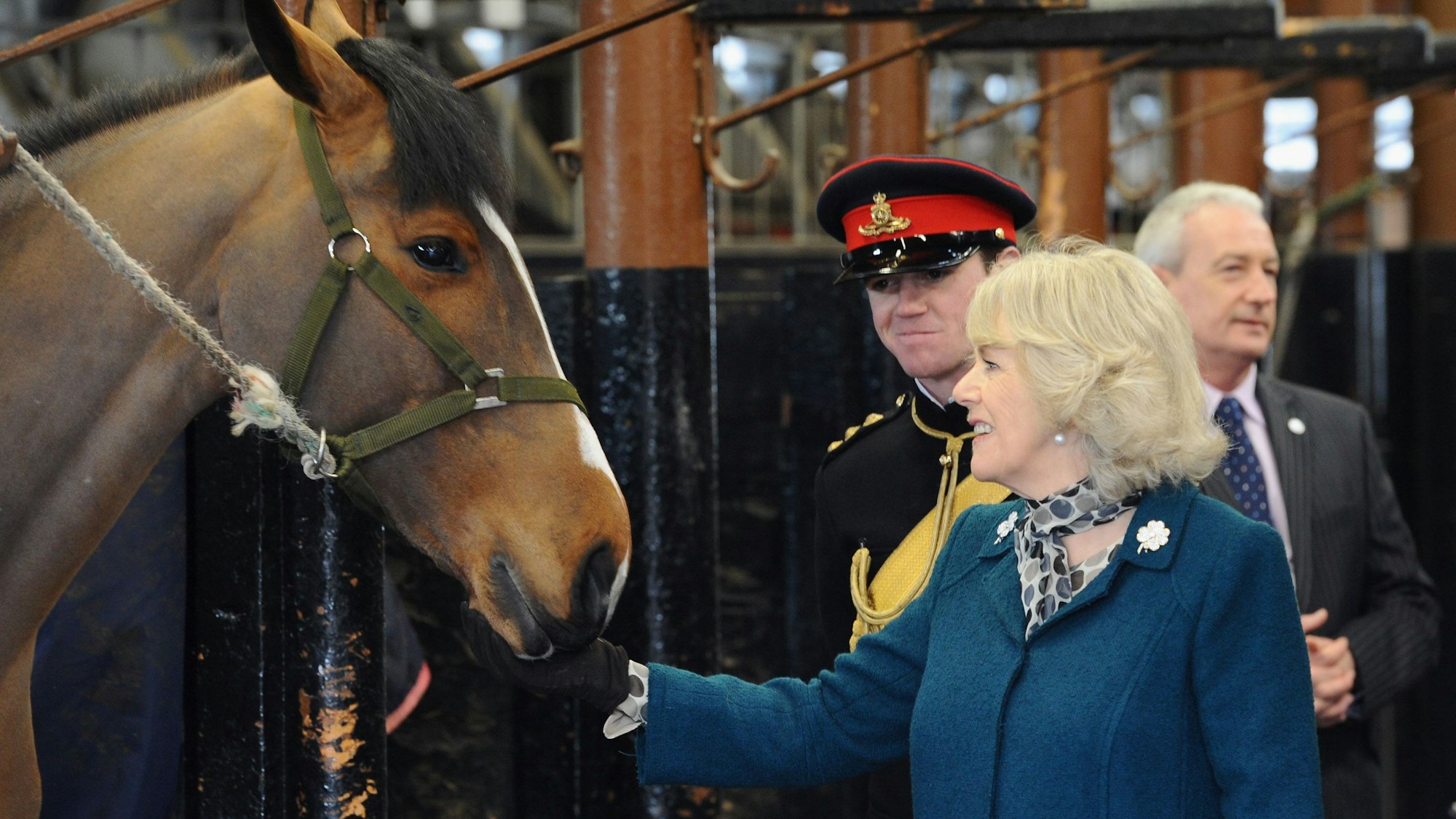 Camilla bei einem Besuch im Pferdestall. Sie streichelt ein Pferd. Ein Wachmann in Uniform schaut ihr dabei zu.