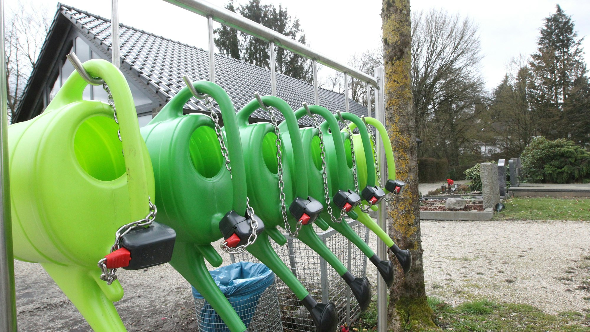 Gießkannen hängen an Stahlketten auf einem Friedhof.