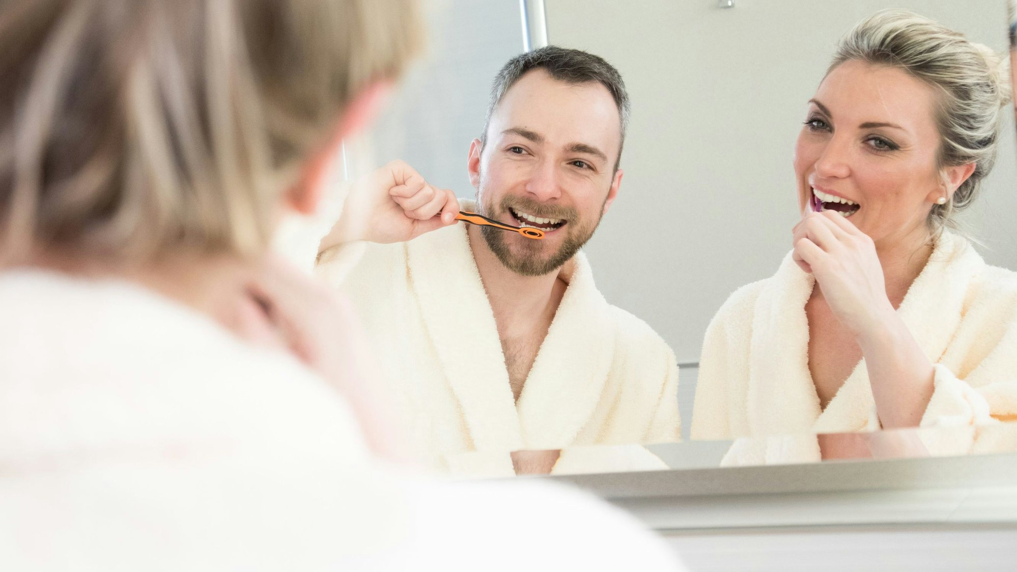 Eine Frau un ein Mann stehen im Bademantel vor dem Waschbecken, im Spiegel sieht man, dass sie sich die Zähne putzen.