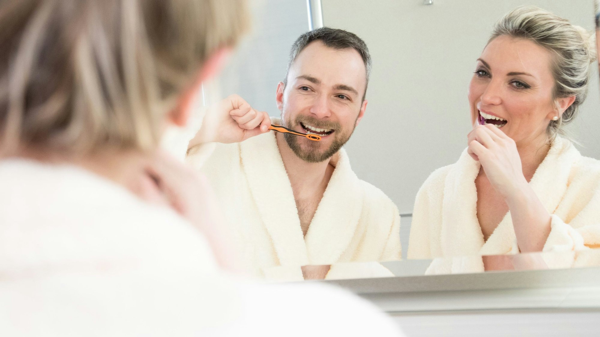 Eine Frau un ein Mann stehen im Bademantel vor dem Waschbecken, im Spiegel sieht man, dass sie sich die Zähne putzen.