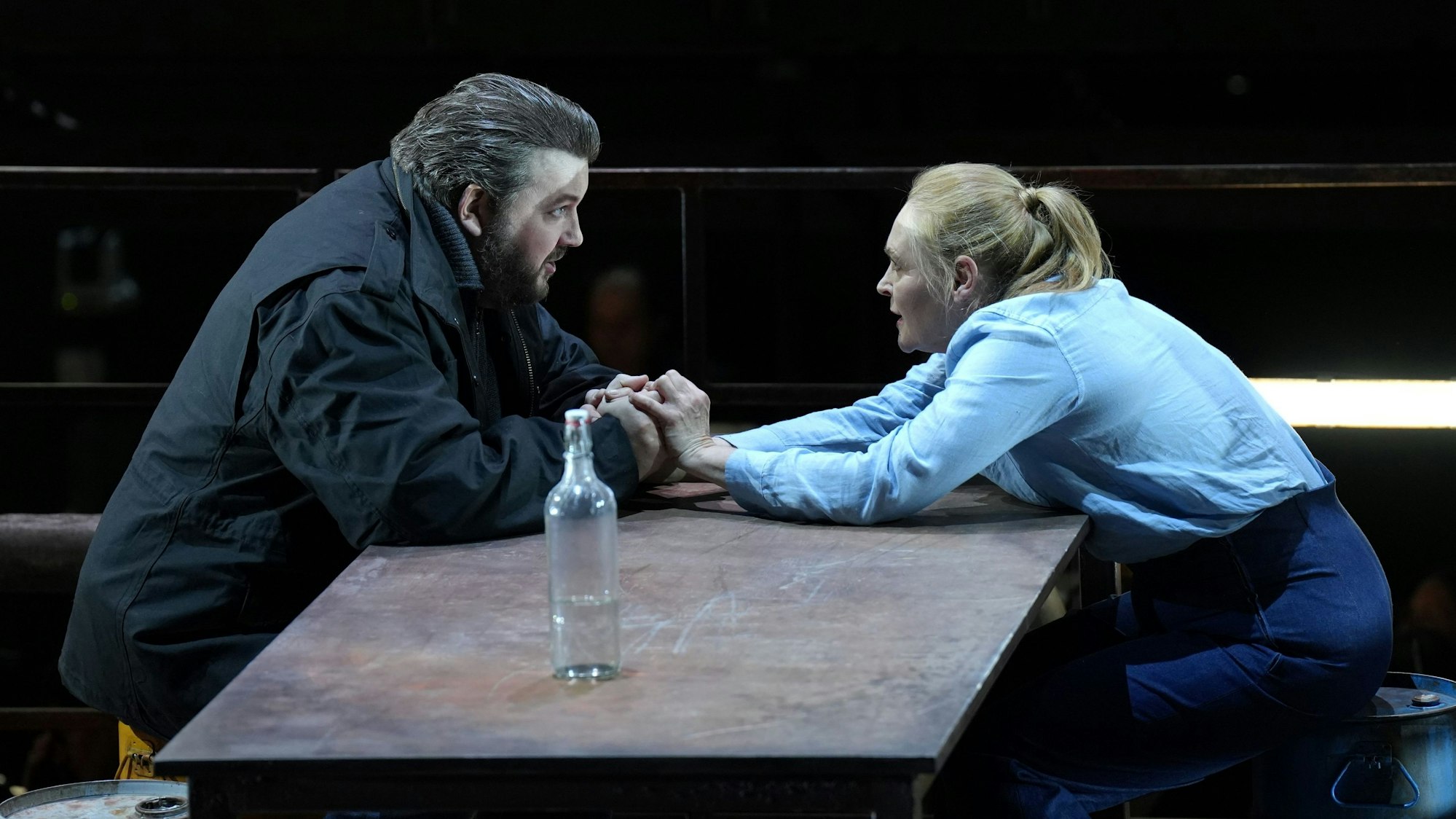 James Rutherford und Ingela Brimberg sitzen an einem Tisch und halten sich die Hände. Eine Glasflasche liegt auf dem Tisch.