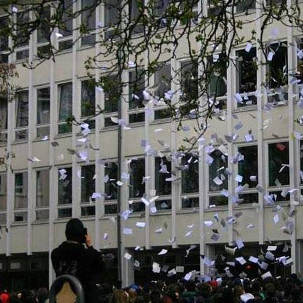 Tausende bedruckte Blätter fliegen aus Fenstern eines Schulgebäudes. Vor dem Gebäude steht eine große Gruppe Schüler.