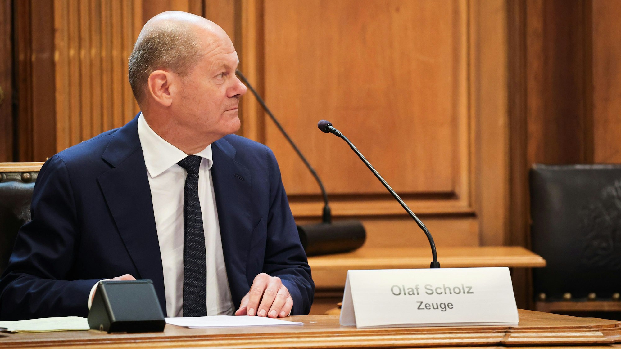 Bundeskanzler Olaf Scholz (SPD) sitzt auf seinem Platz auf der Senatsbank mit einem Plastikschild mit dem Text ·Olaf Scholz Zeuge· vor Beginn einer Sitzung des Parlamentarischen Untersuchungsausschusses ·Cum-Ex· im Plenarsaal der Hamburgischen Bürgerschaft im Rathaus.