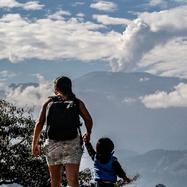 Es ist eine Mutter mit ihrem Kind an der Hand zu sehen. Im Hintrgrund steht der Vulkan.