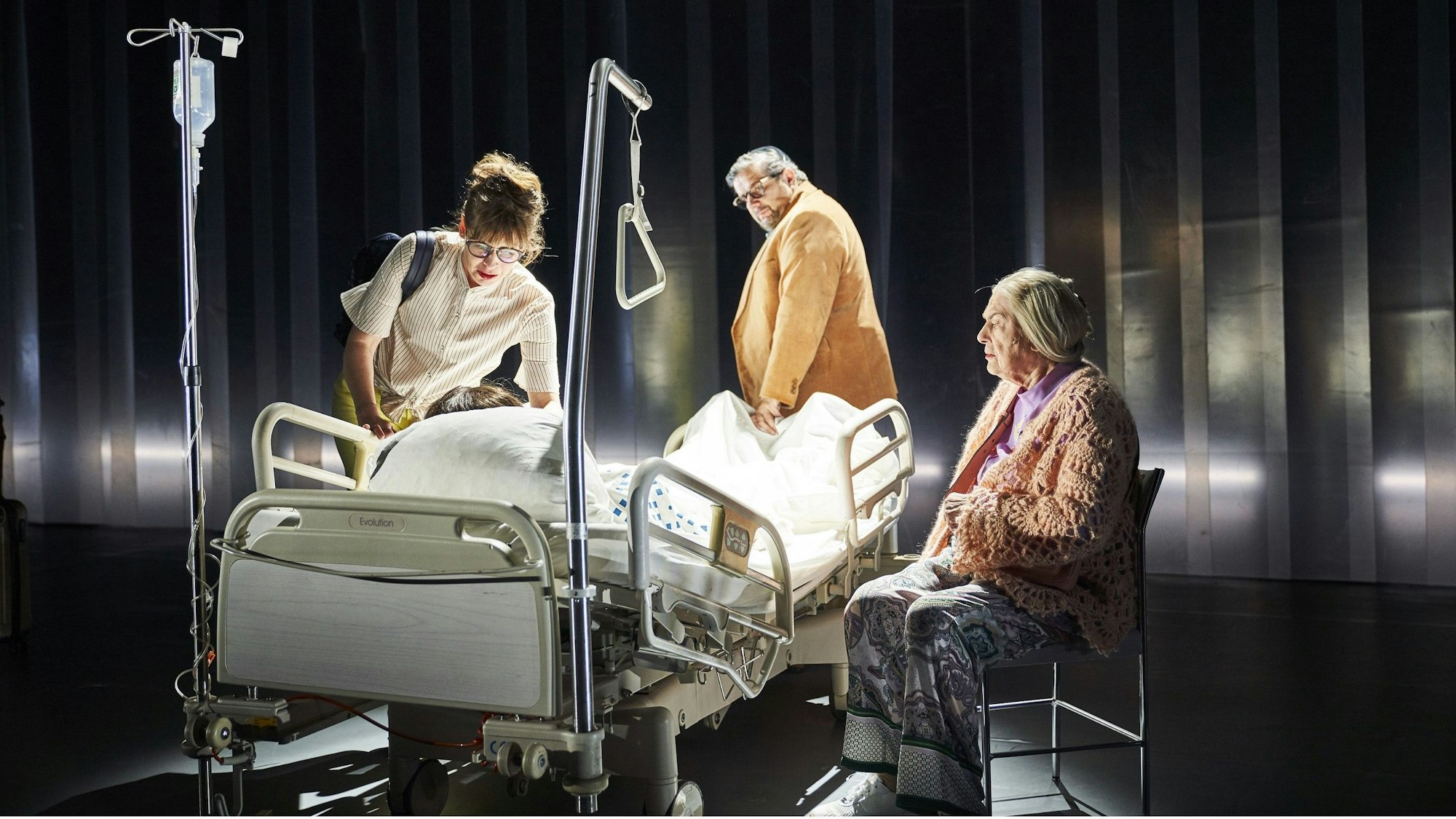 Drei Menschen stehen und sitzen an einem Krankenhausbett.