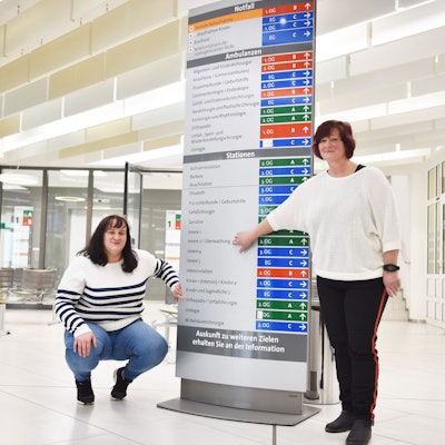Zwei Frauen zeigen auf einen Wegweiser, der zu den Stationen in einem Krankenhaus führt.