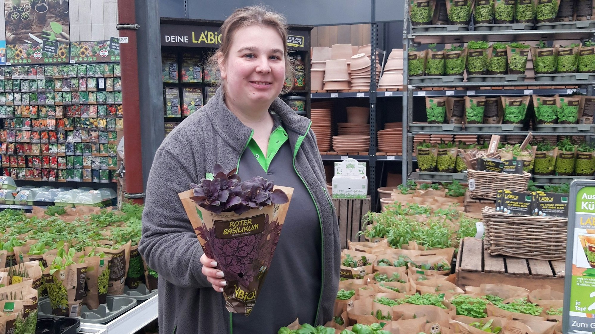 Eine Gartenexpertin in einem Blumenladen hält grinsend einen Blumentopf vor sich.