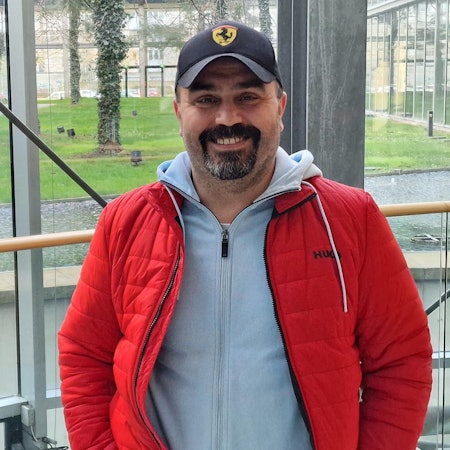 Canan Karadag, in roter Steppjacke und mit Ferrari-Kappe, steht im Neven DuMont Haus in Köln und lächelt in die Kamera.