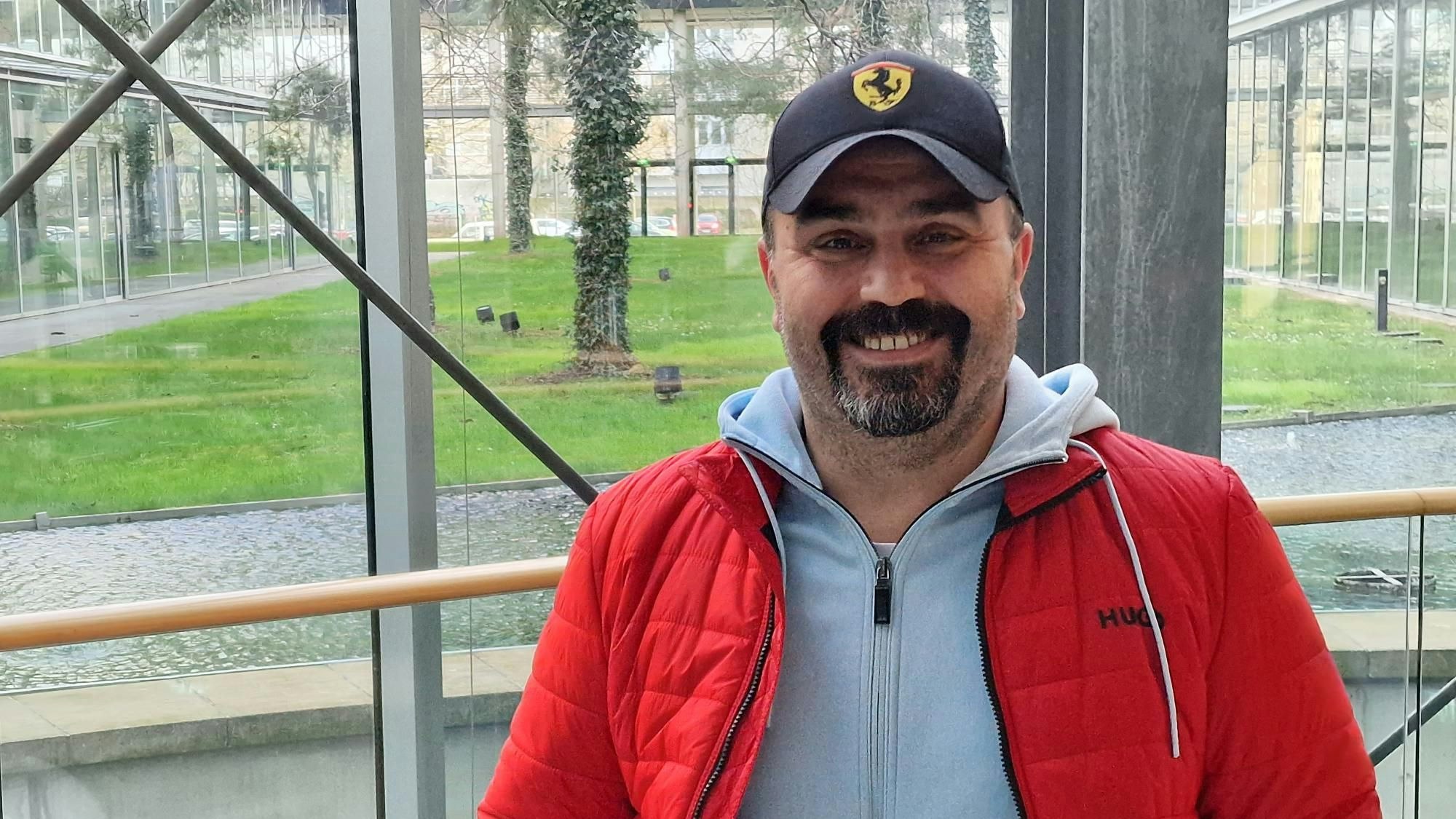 Canan Karadag, in roter Steppjacke und mit Ferrari-Kappe, steht im Neven DuMont Haus in Köln und lächelt in die Kamera.