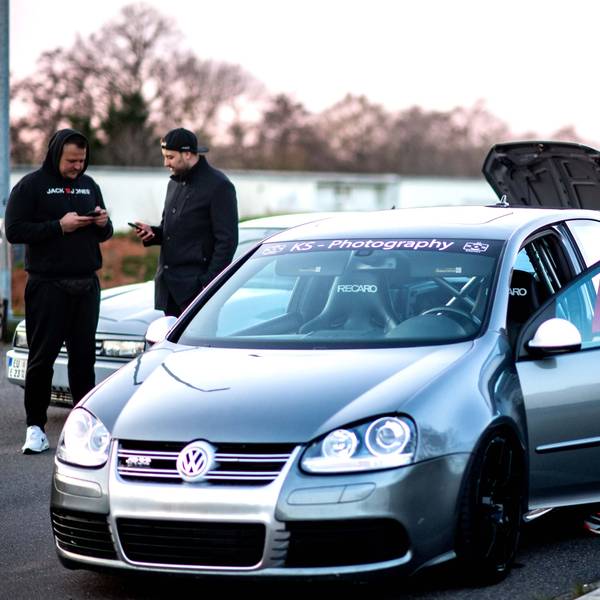 Michelle Engel steht an ihrem Golf R32. Im Hintergrund schauen zwei Männer auf ihr Handy.